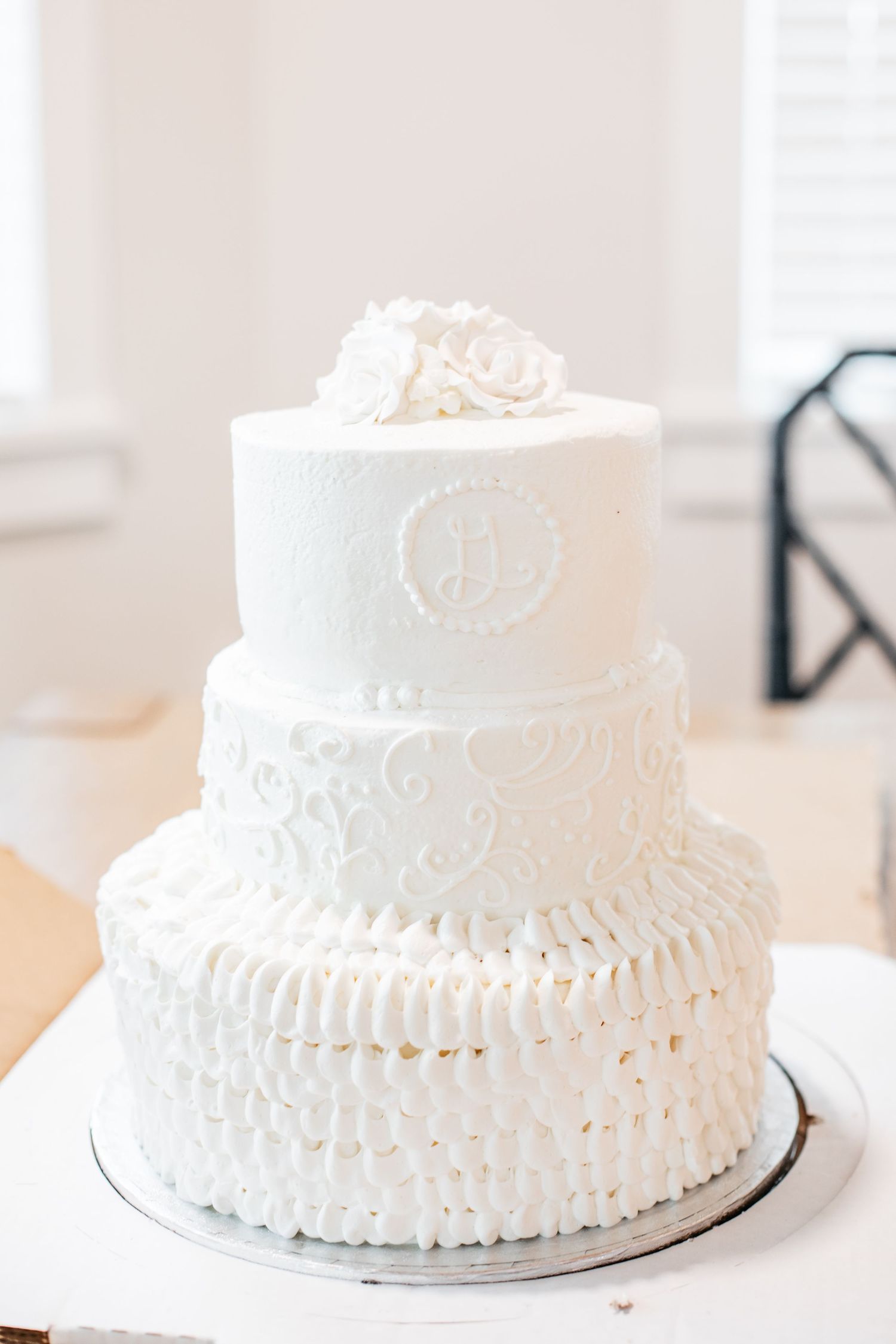 Elegant three-tier white wedding cake with ruffles, scrollwork, monogram, and floral topper.
