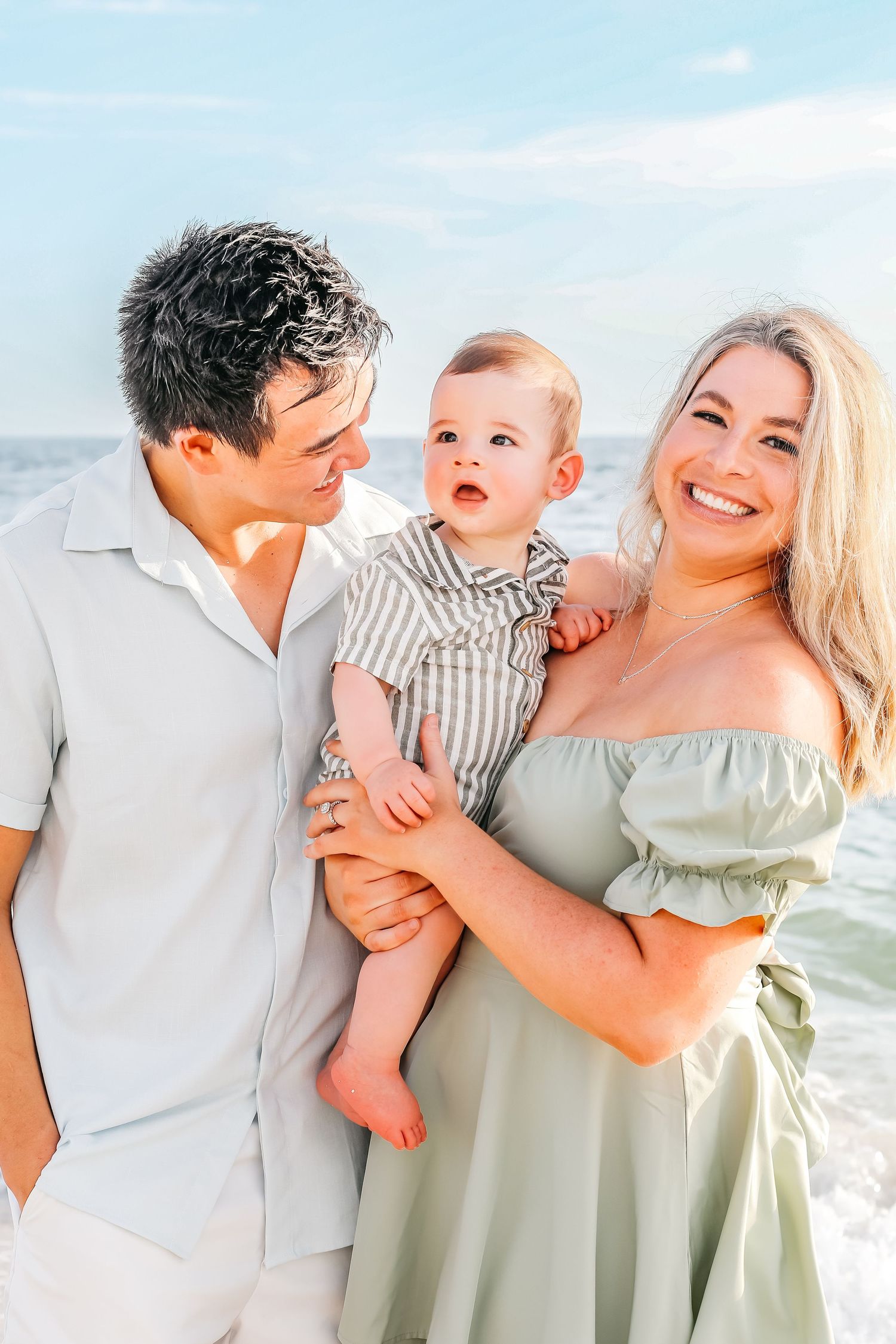Joyful family beach portraits with baby wearing coordinated sage green and white outfits.