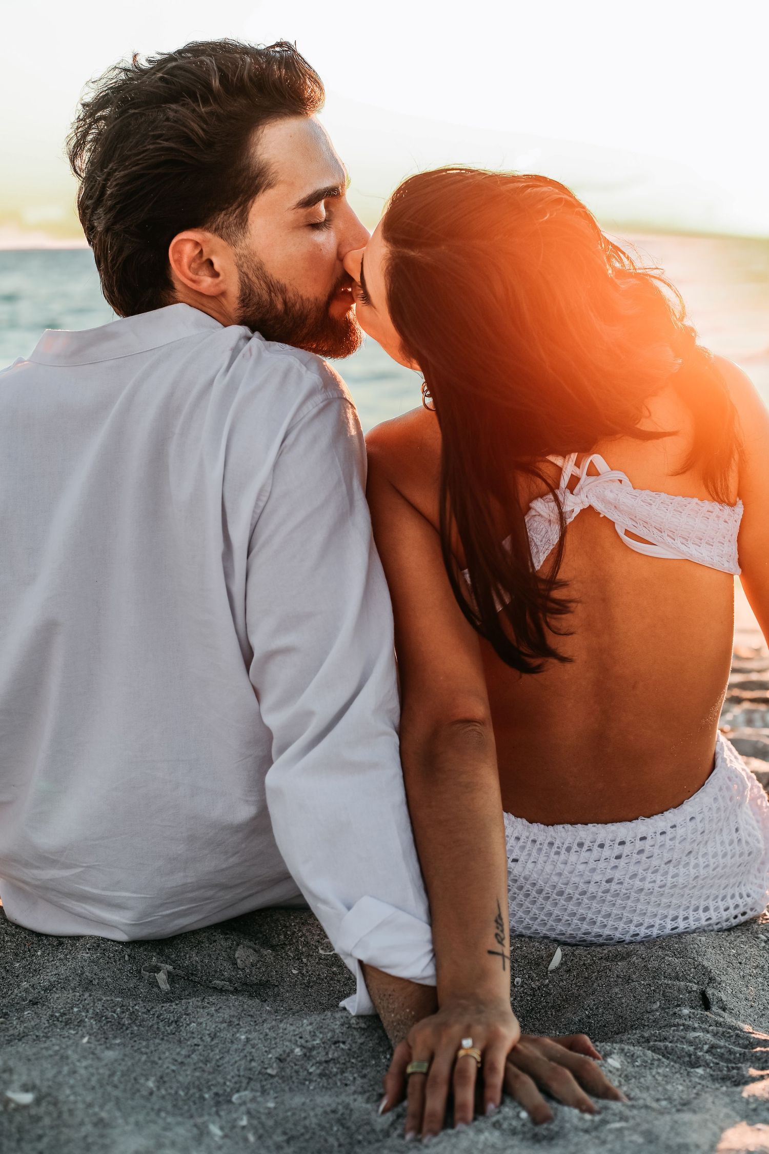 A couple in casual attire sits close together on a beach during sunset, sharing an intimate moment.