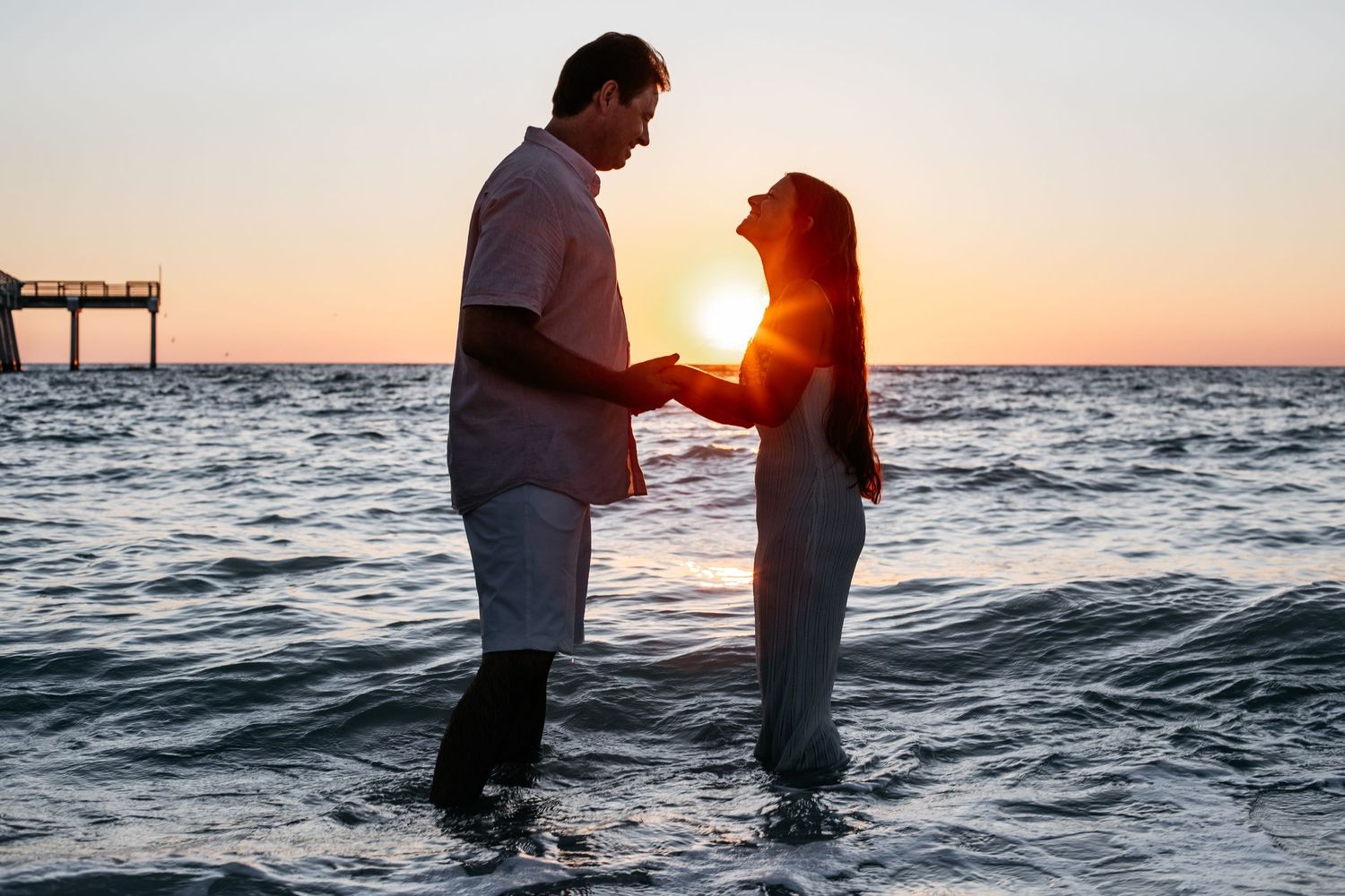 Silhouetted couple stands in ocean water during golden sunset creating romantic beach atmosphere.