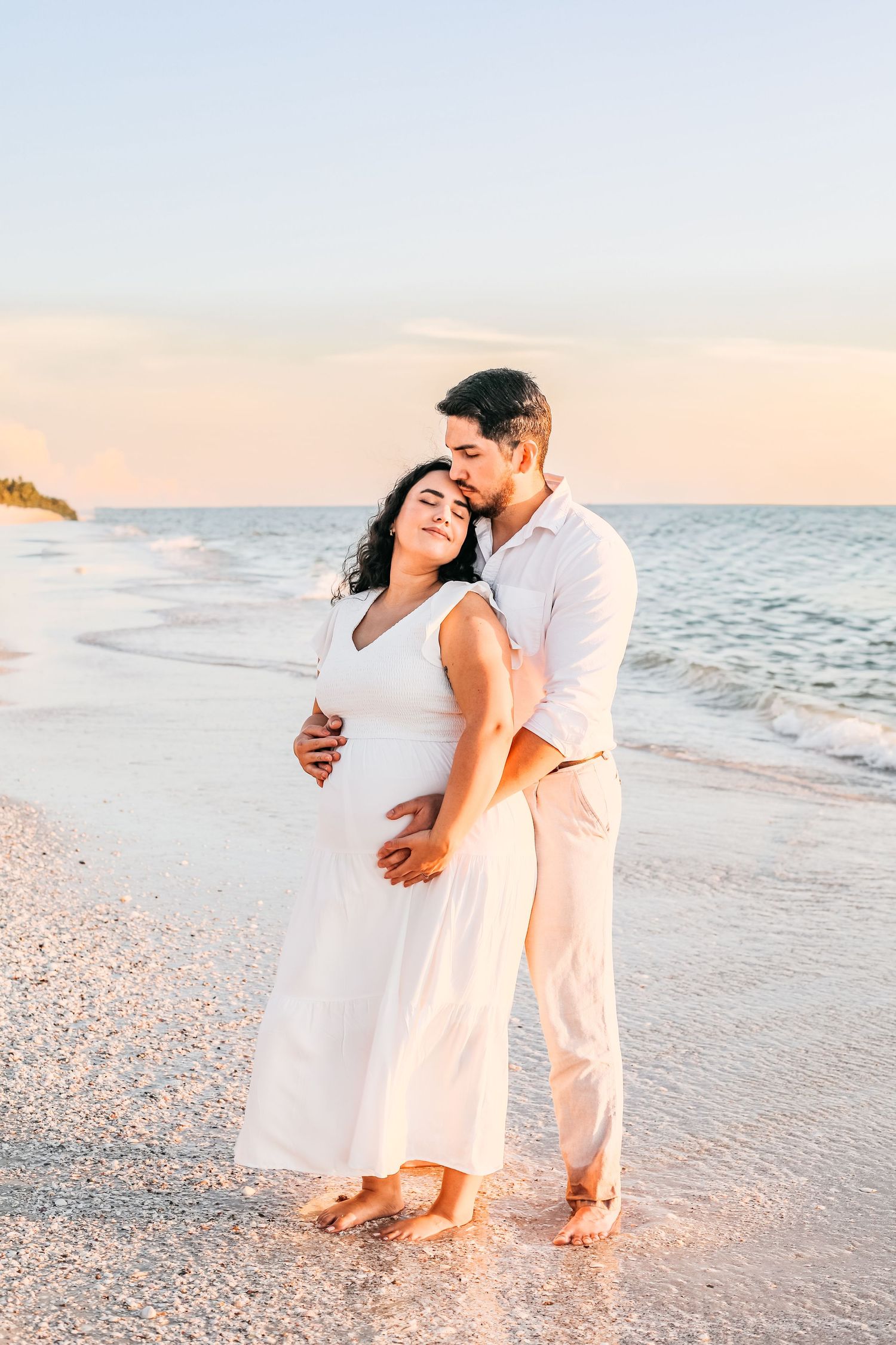 A romantic couple in white outfits embraces on a sandy beach at sunset with ocean waves in the background.