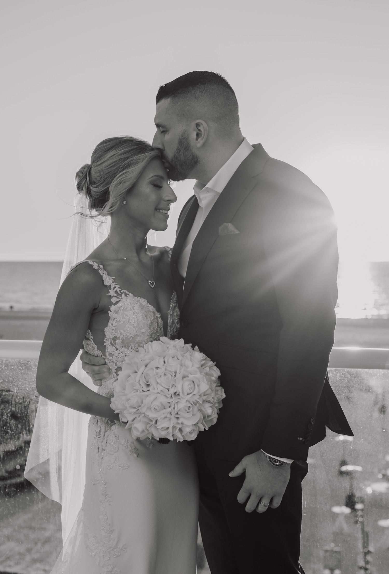 B&W of a groom kissing his bride with the sunset shining through.
