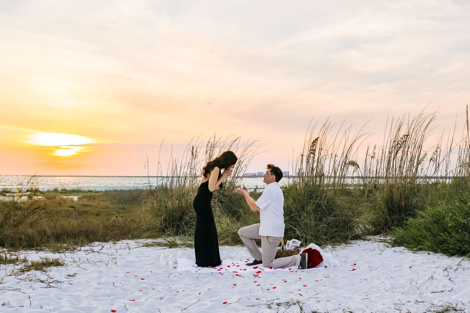 Romantic beach proposal at sunset, man on one knee with rose petals on white sand dunes.