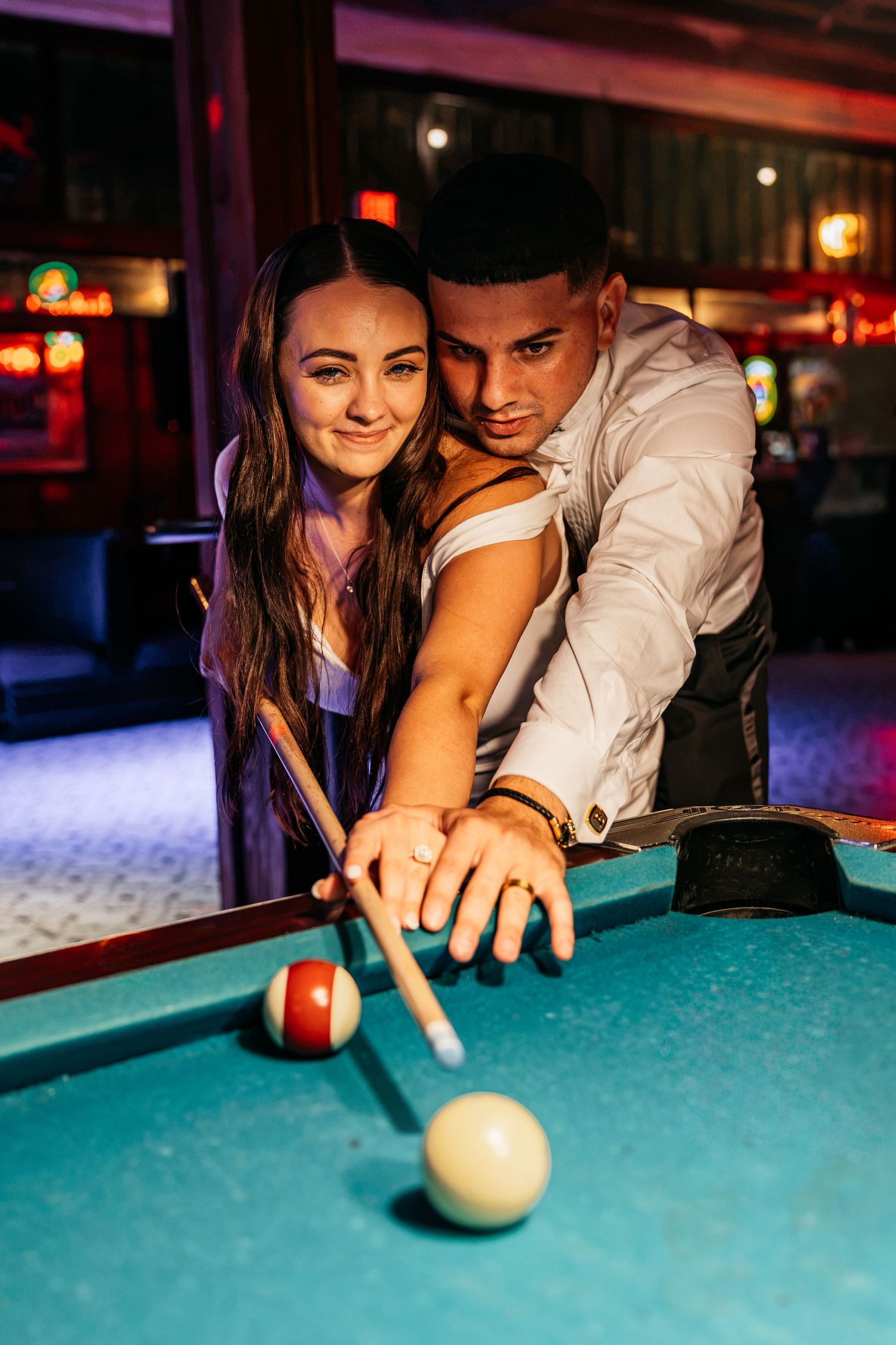 Couple playing pool together at a bar, woman smiling as man guides her shot on a green billiard table.