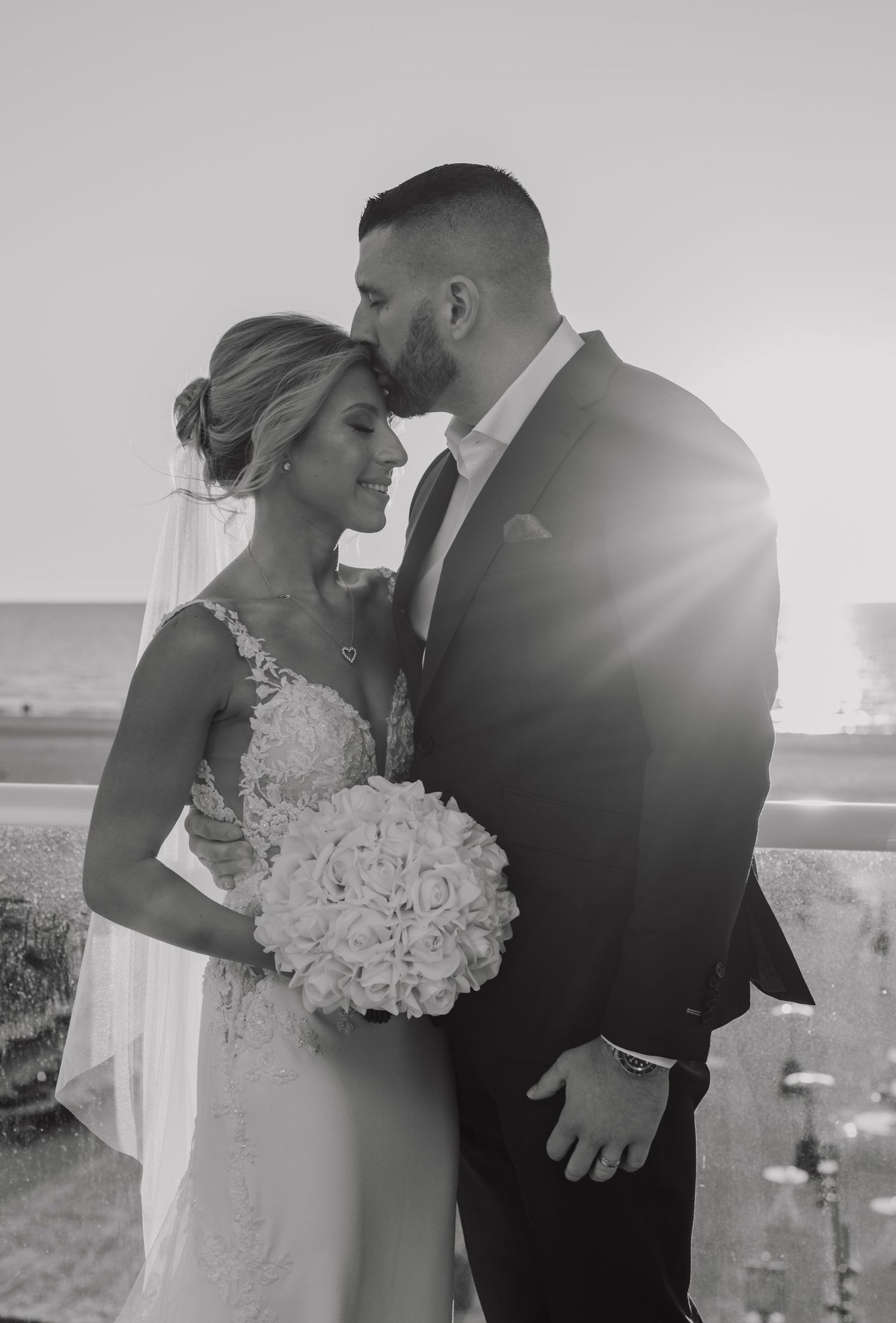 Black and white wedding photo of bride holding bouquet and groom kissing her forehead at sunset by the ocean.