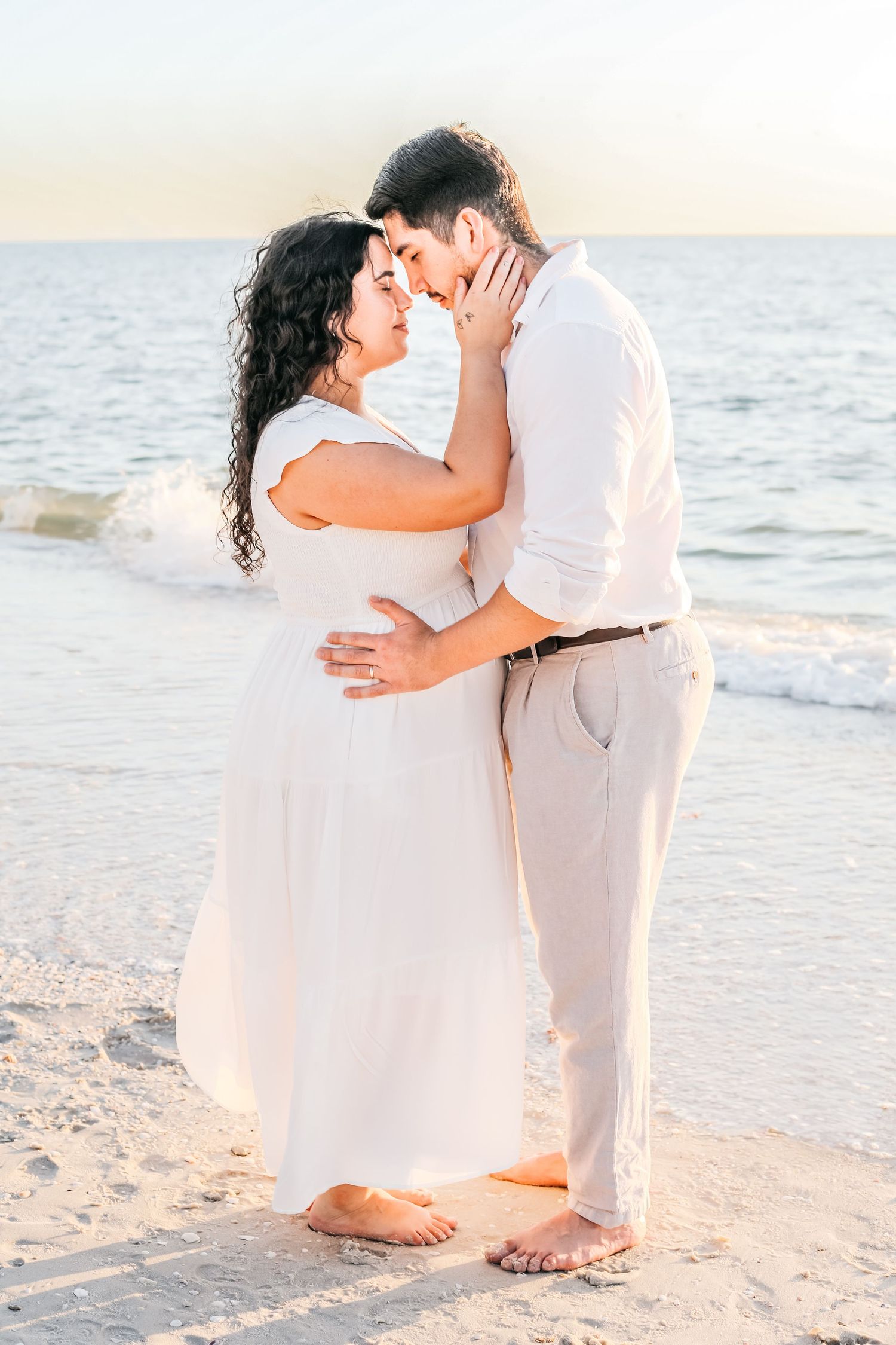 A couple shares intimate moments on a pristine beach with gentle waves in the background at dusk.