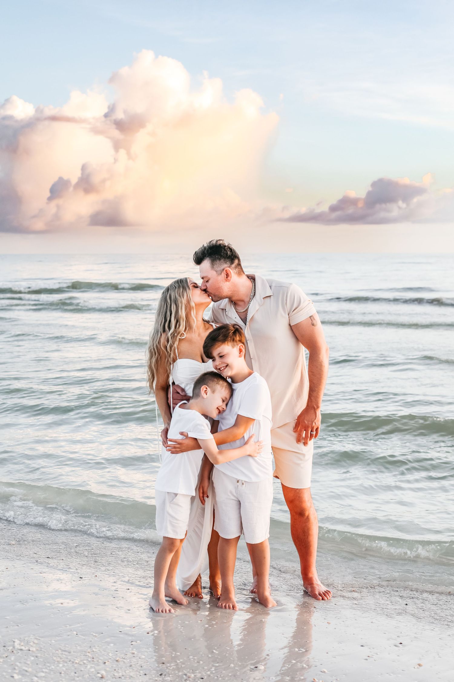 A family wearing white outfits shares tender moments together on a beach at sunset with ocean waves in the background.