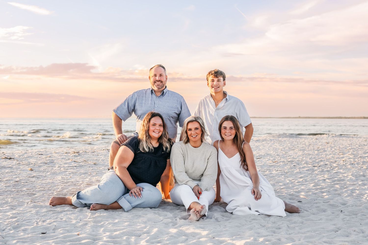 A group wearing white and blue casual clothing poses together on a sandy beach at sunset.
