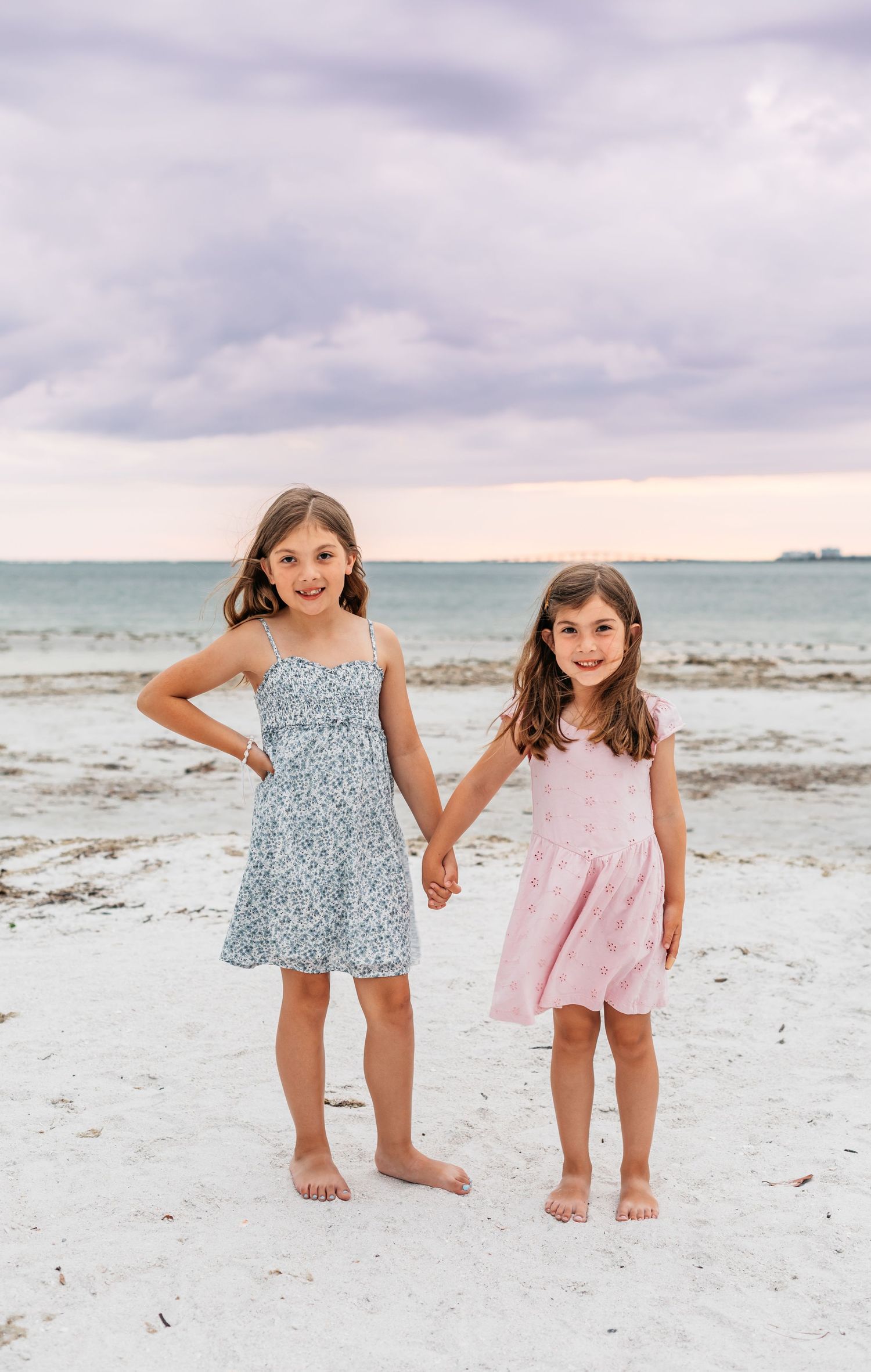 Two young girls in summer dresses hold hands on a white sand beach with calm ocean waters behind them.