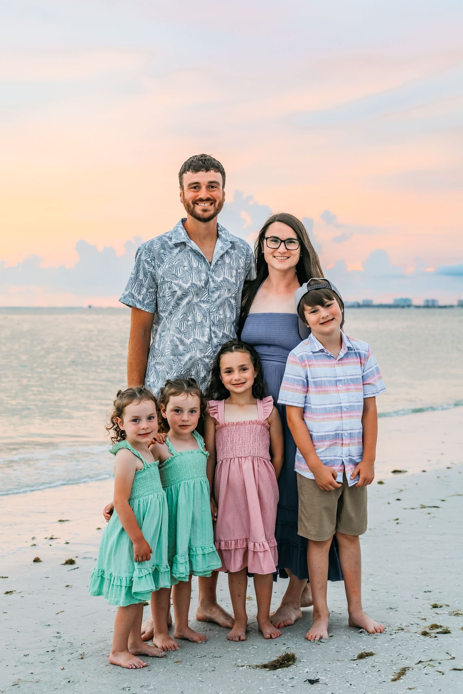Family of six posing on a beach at sunset, parents standing behind four young children in colorful summer outfits.