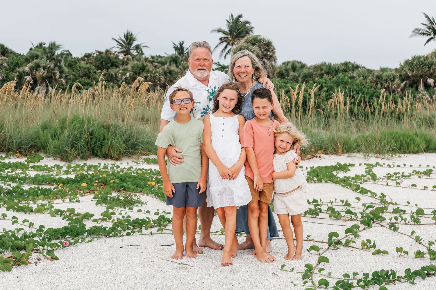 A group stands together on a sandy beach with tall grass and palm trees in the background.