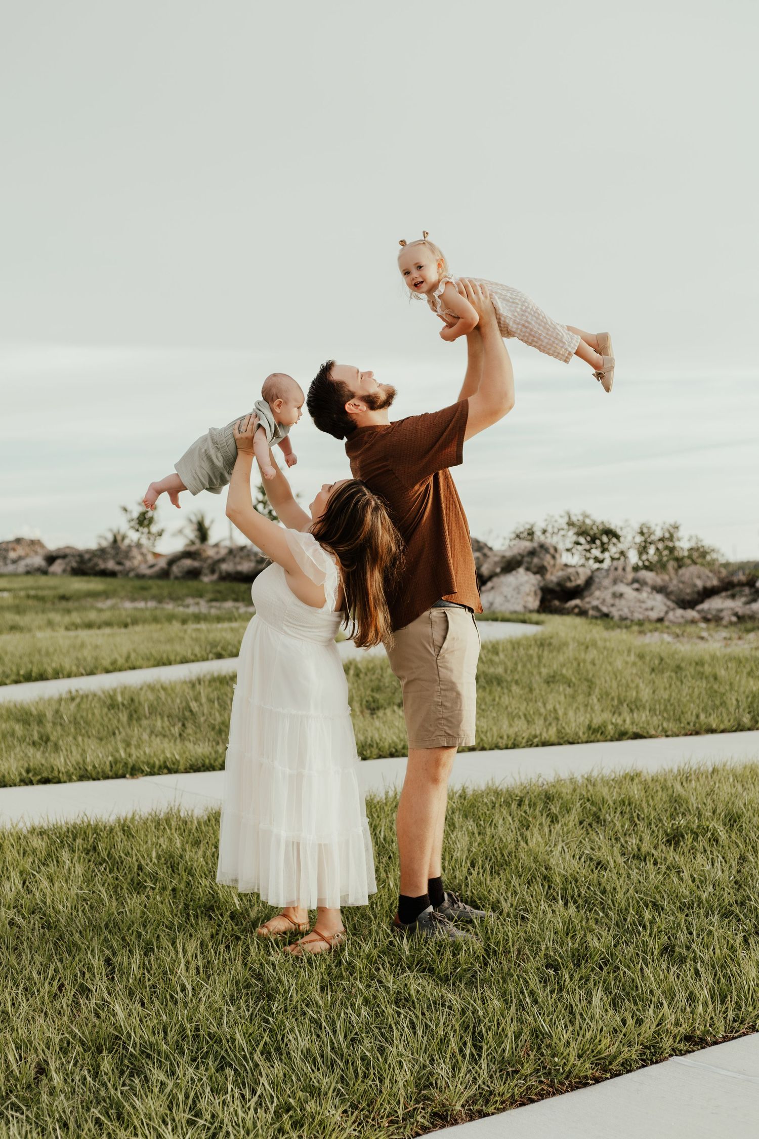 A joyful family outdoors, parents lifting two young children in the air on a green lawn at dusk.