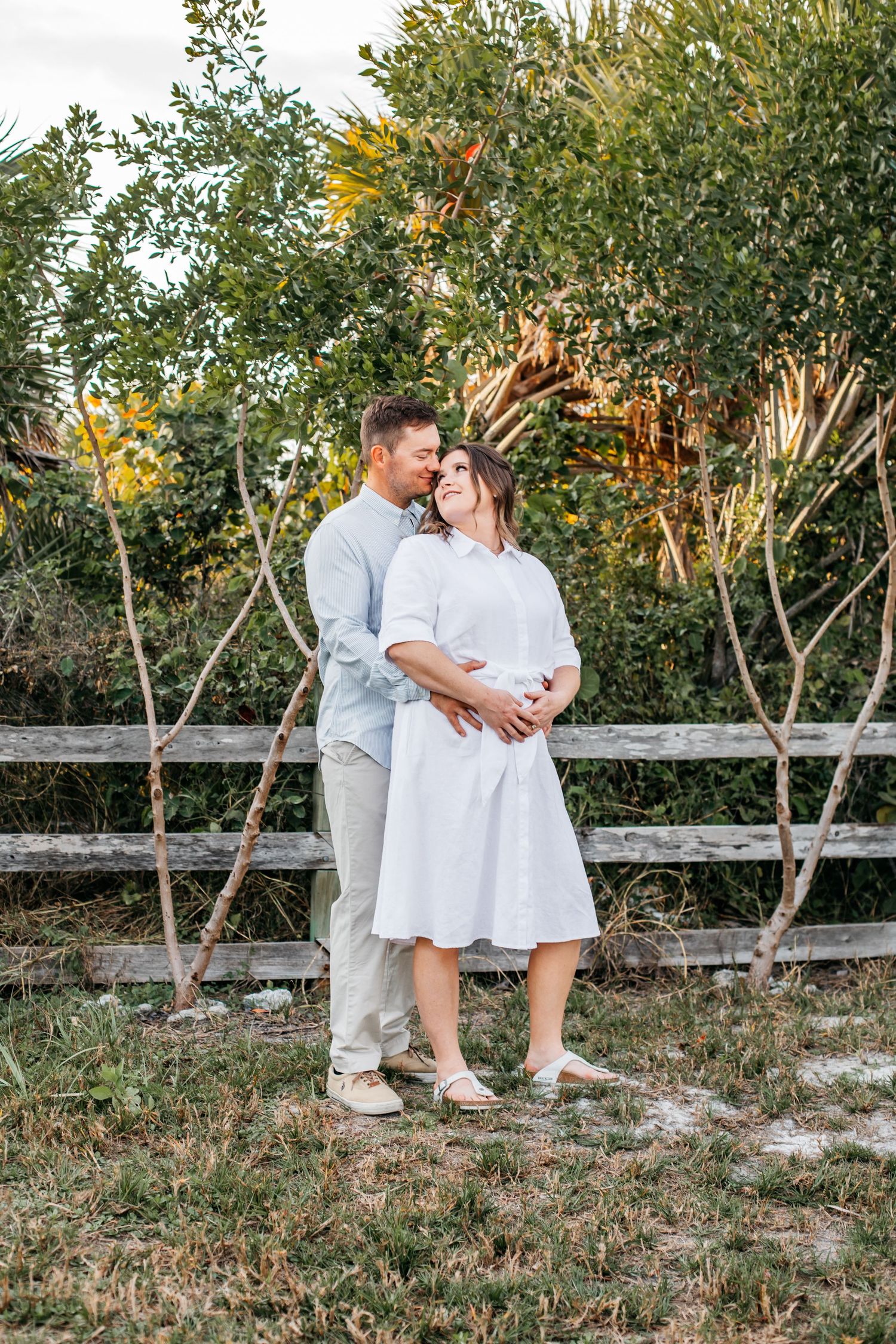 A couple embraces outdoors near a wooden fence, the woman in a white dress and man in light blue, surrounded by lush greenery.