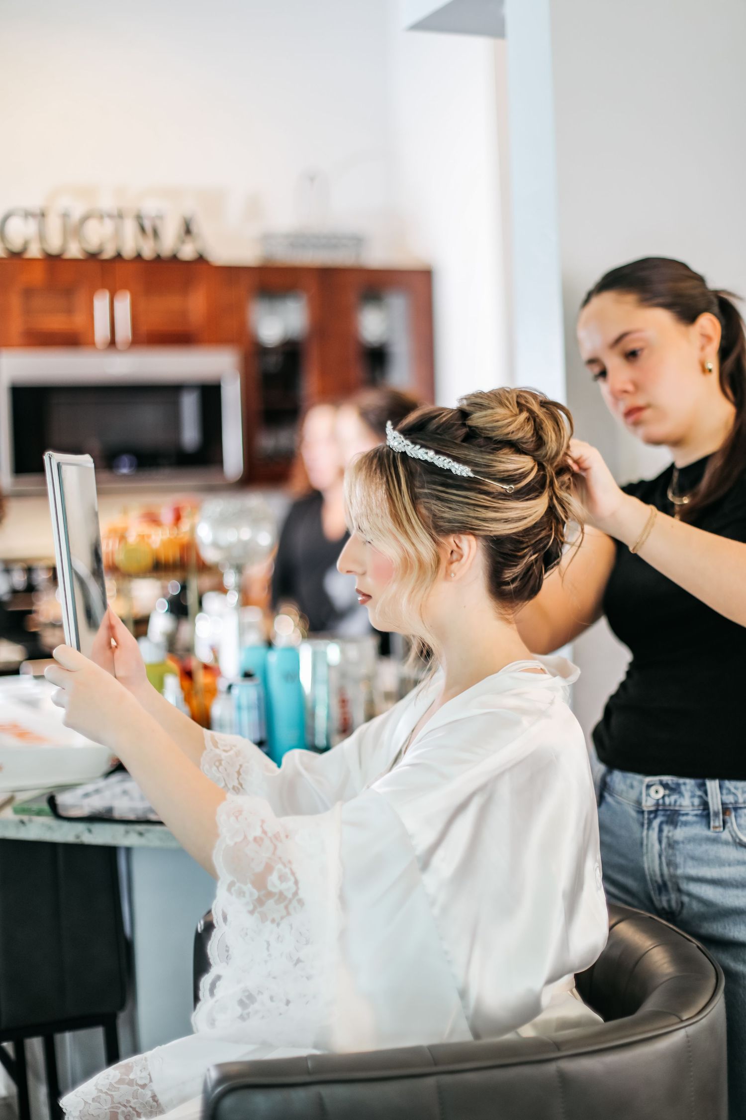 Bride in white robe checks mirror while hairstylist arranges updo with crystal headband in bright salon.