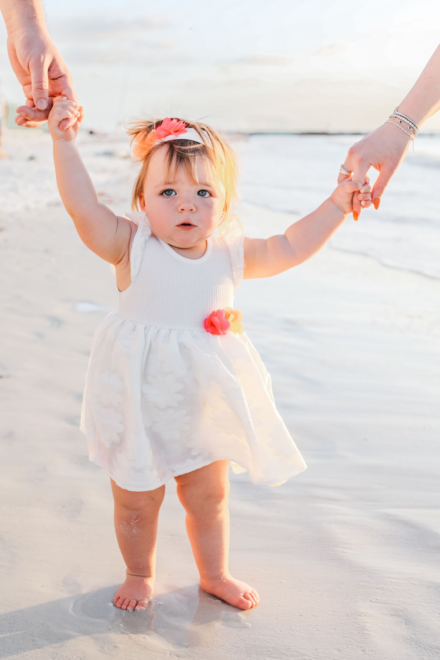 A small child in a white dress takes first steps on a sandy beach while being supported by adults' hands.