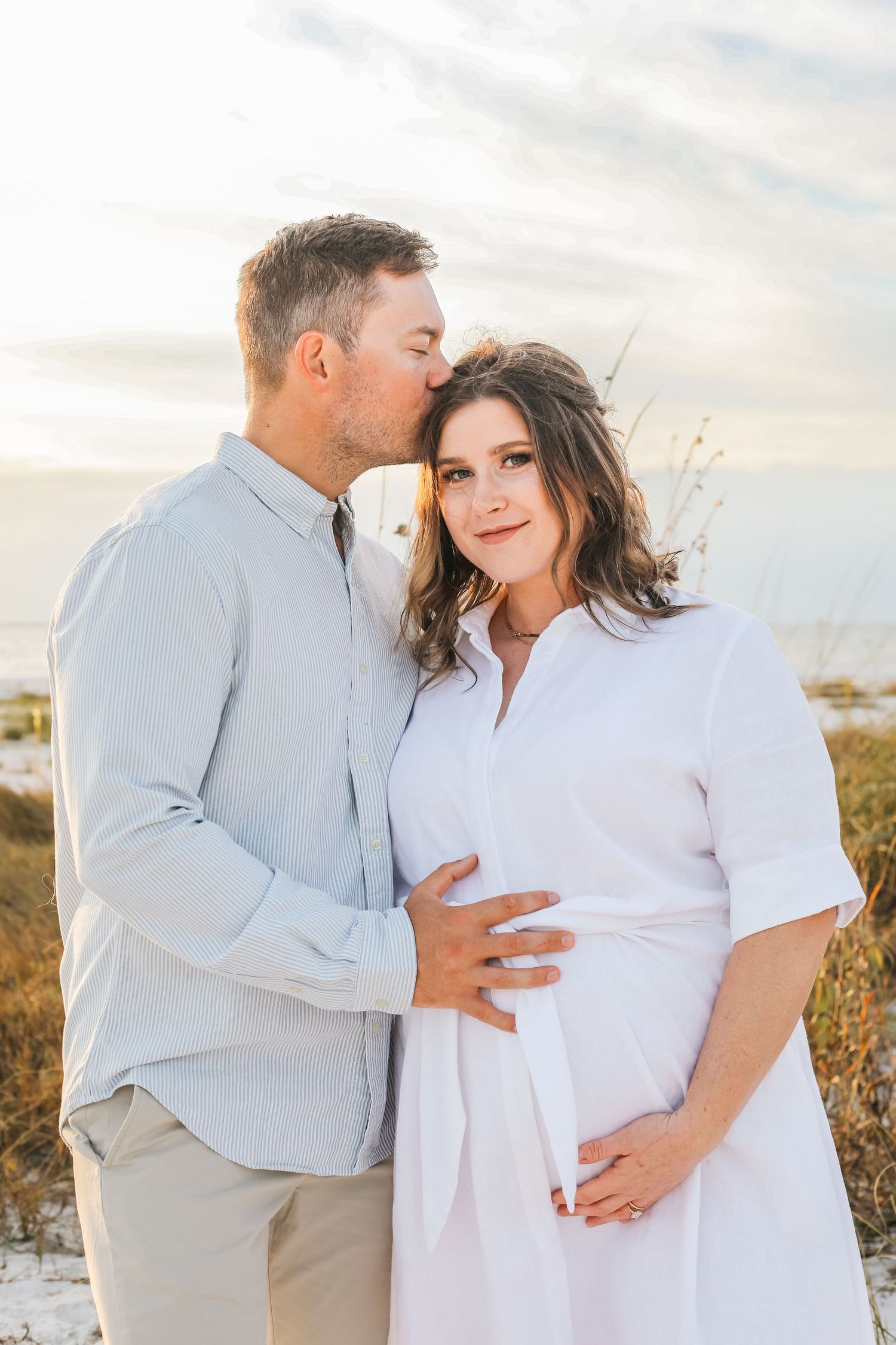 A man kisses a pregnant woman on the forehead at a beach during golden hour, both smiling warmly.