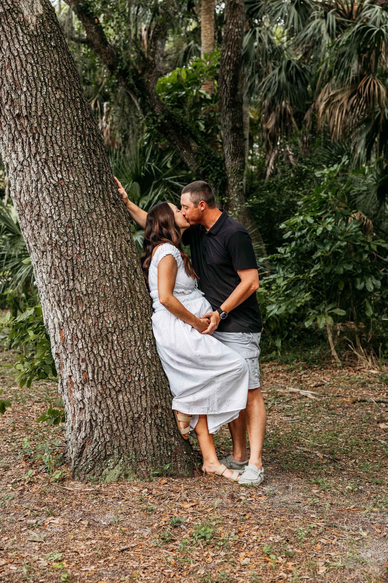 A couple shares a romantic kiss leaning against a large tree trunk in a lush green outdoor setting.