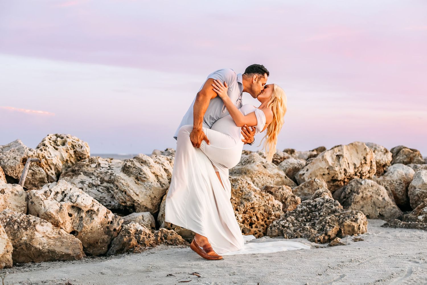 A couple in white wedding attire embraces on a rocky beach at sunset with warm golden lighting.