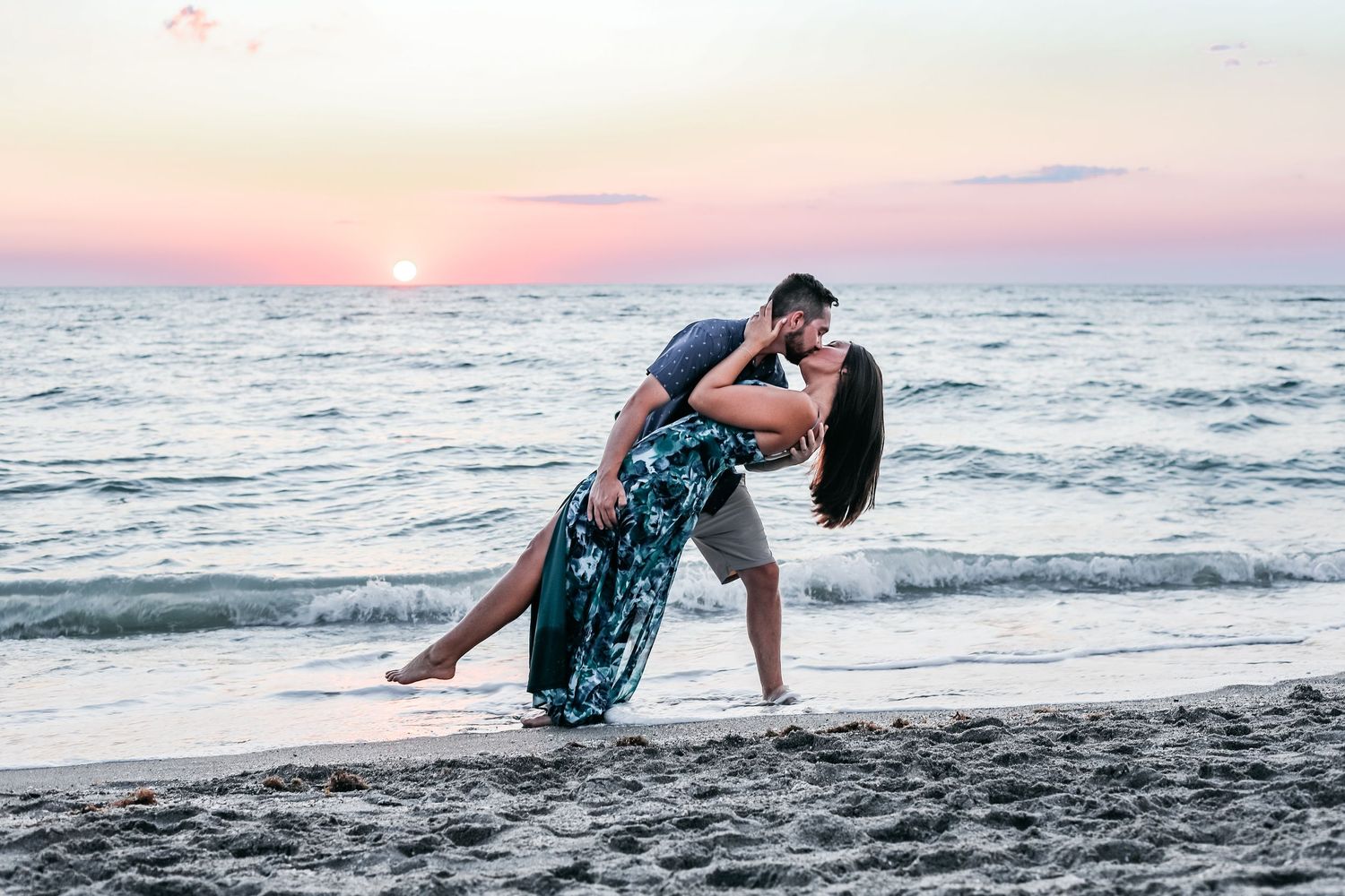 A romantic couple embraces in a dip pose on the beach at sunset with ocean waves in the background.