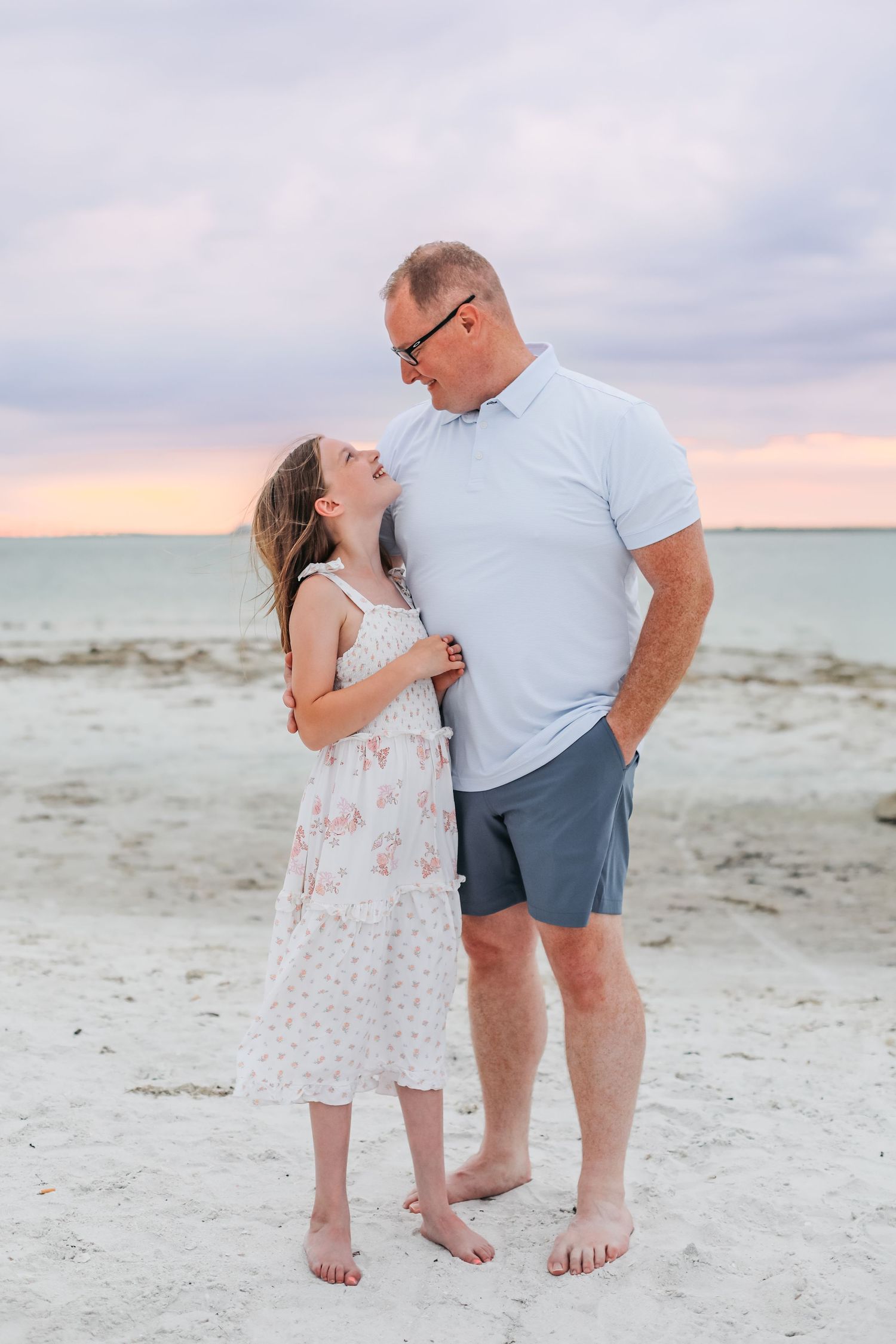 A father and daughter share a sweet moment on a sandy beach at sunset, with colorful pastel skies in the background.