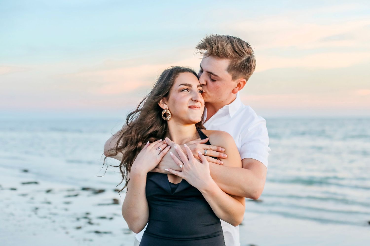 Romantic couple embracing on a beach at sunset, man kissing woman's cheek near ocean waves.