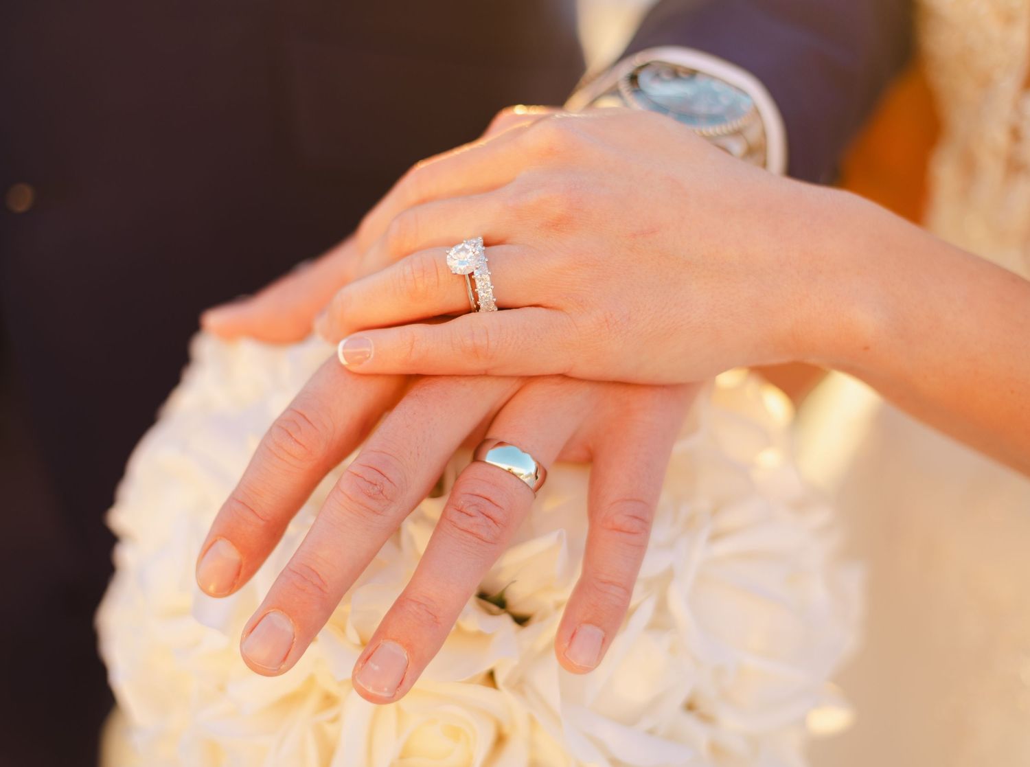 Couple displaying wedding rings on ring fingers, bride holding white roses bouquet, diamond engagement ring visible.