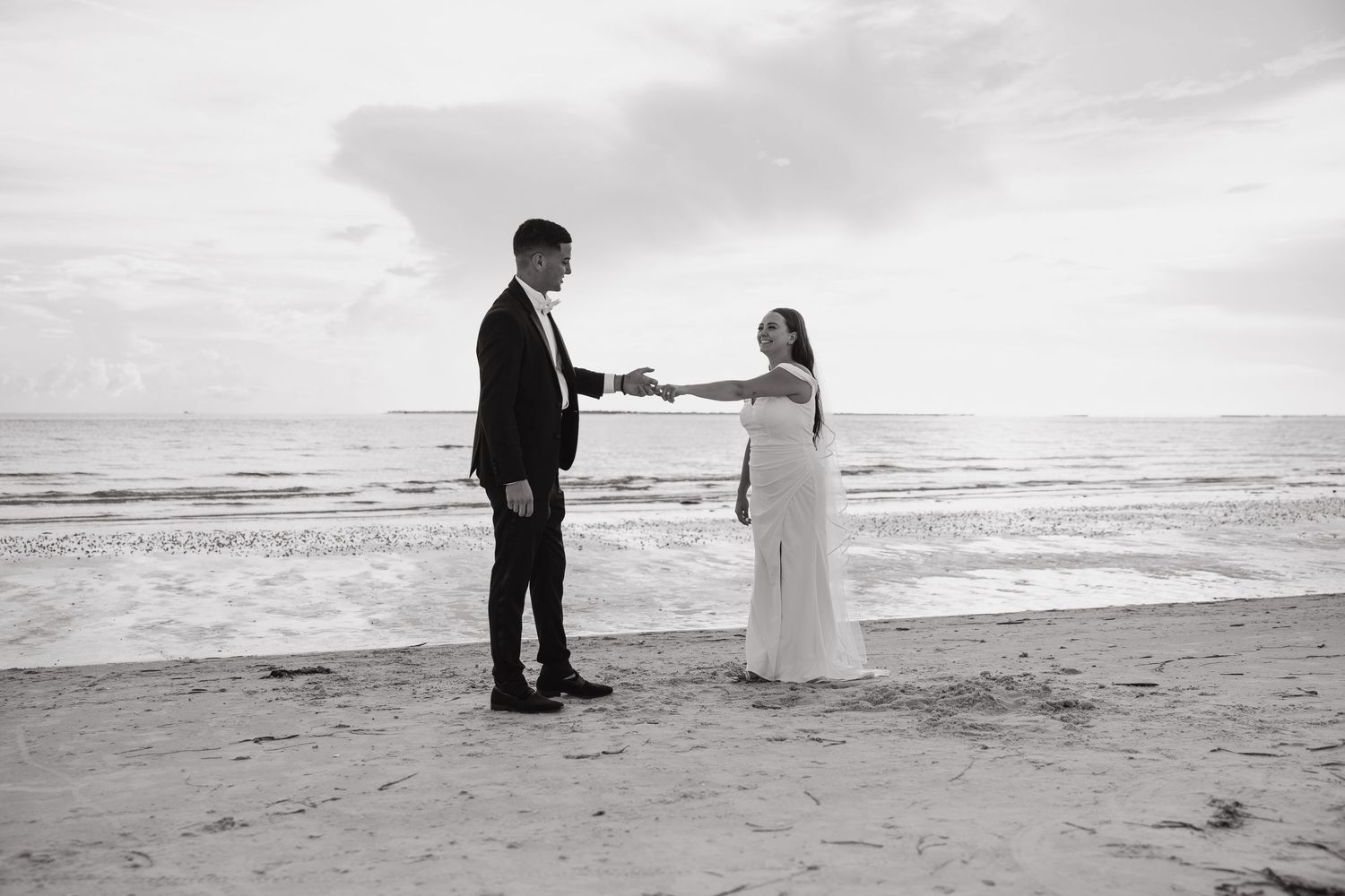 Black and white beach wedding photo of couple in formal attire holding hands by the ocean shore.