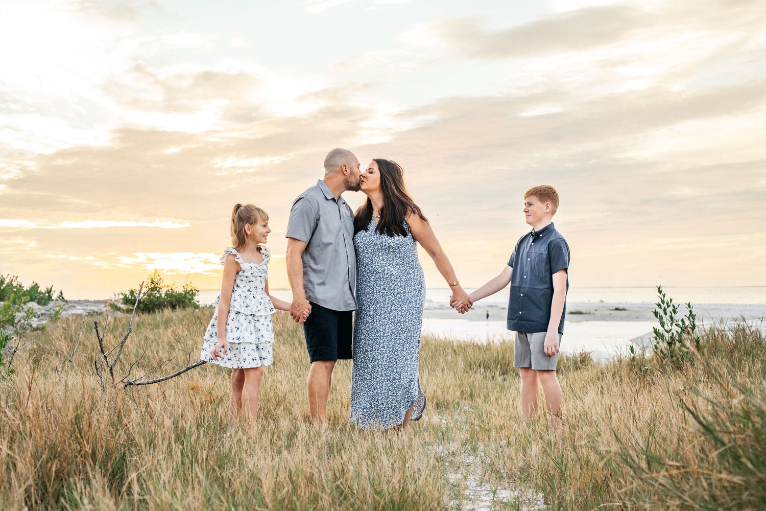 Family of four holding hands at sunset on a grassy beach path, parents kissing while children stand beside them.