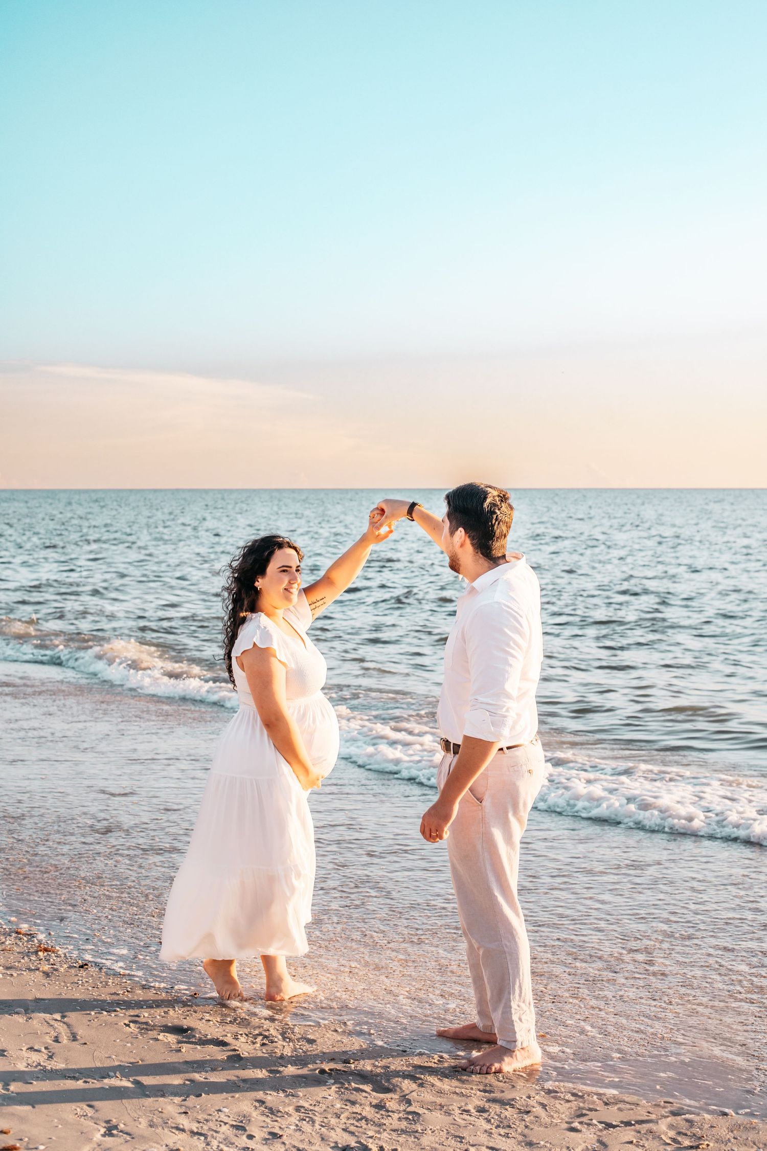 A couple in white attire dances together playfully on a sandy beach at sunset with ocean waves in the background.