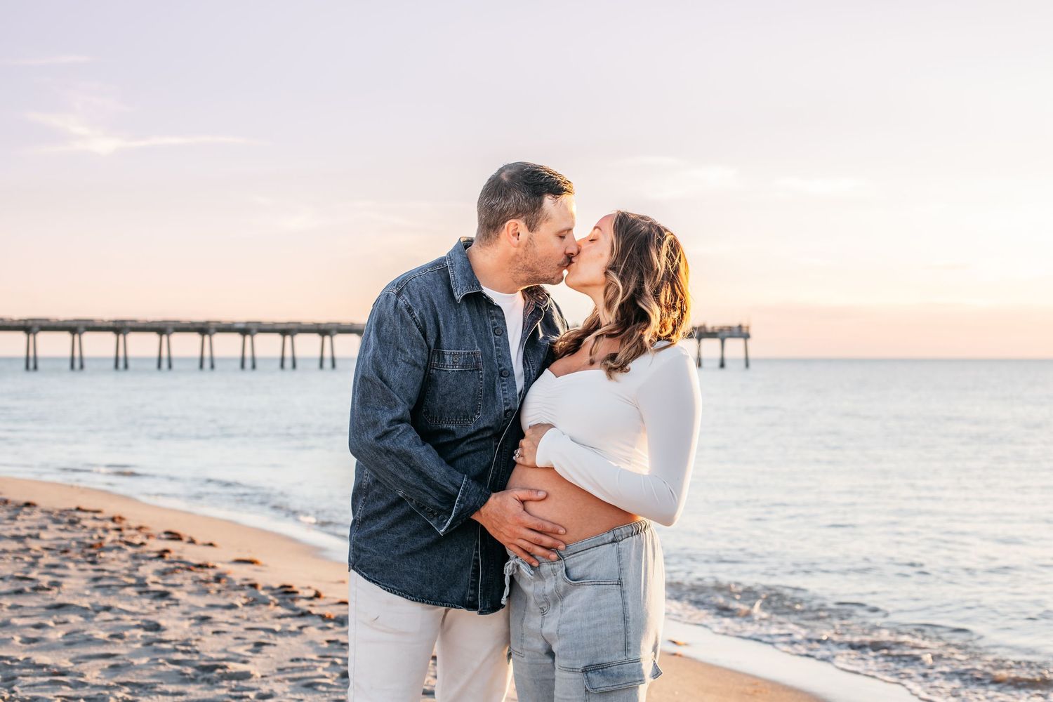 Romantic couple shares intimate moments on a sandy beach at sunset with a pier in the background.