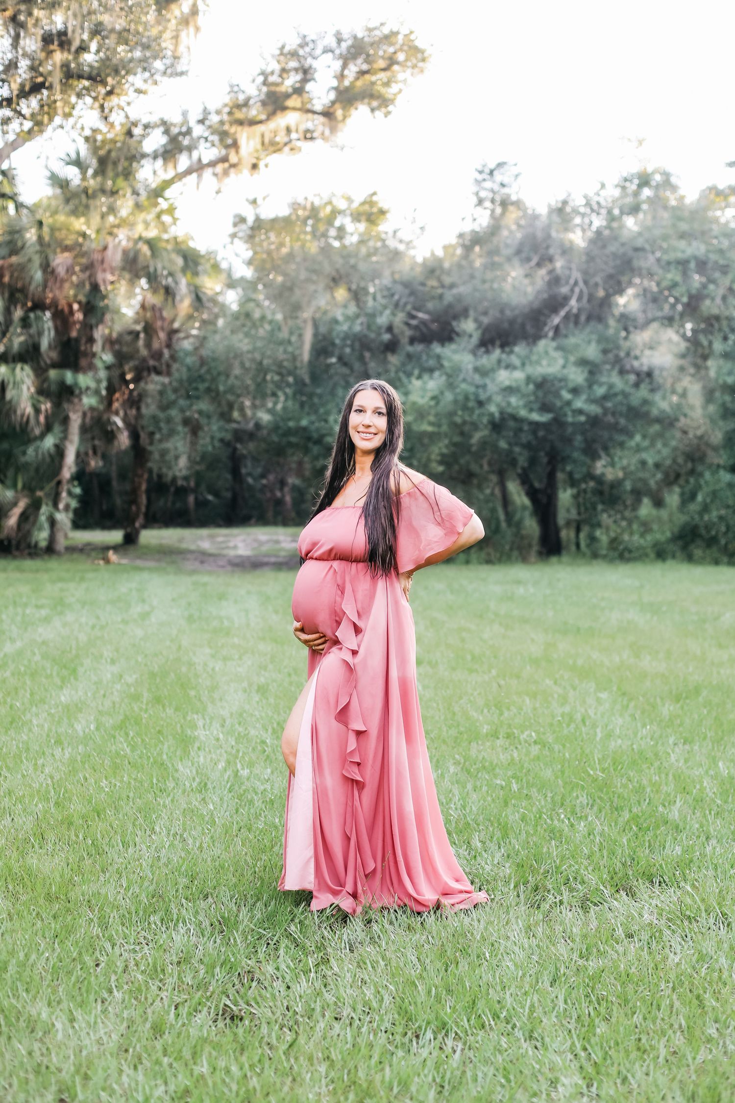 A person in a flowing coral maxi dress twirls joyfully in a sunlit grassy field with trees in the background.