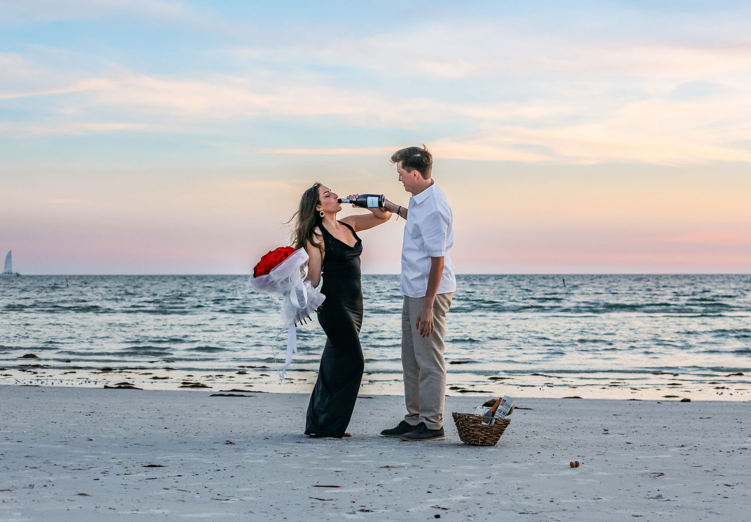 Couple sharing champagne on a beach at sunset with roses and a picnic basket during a romantic proposal.