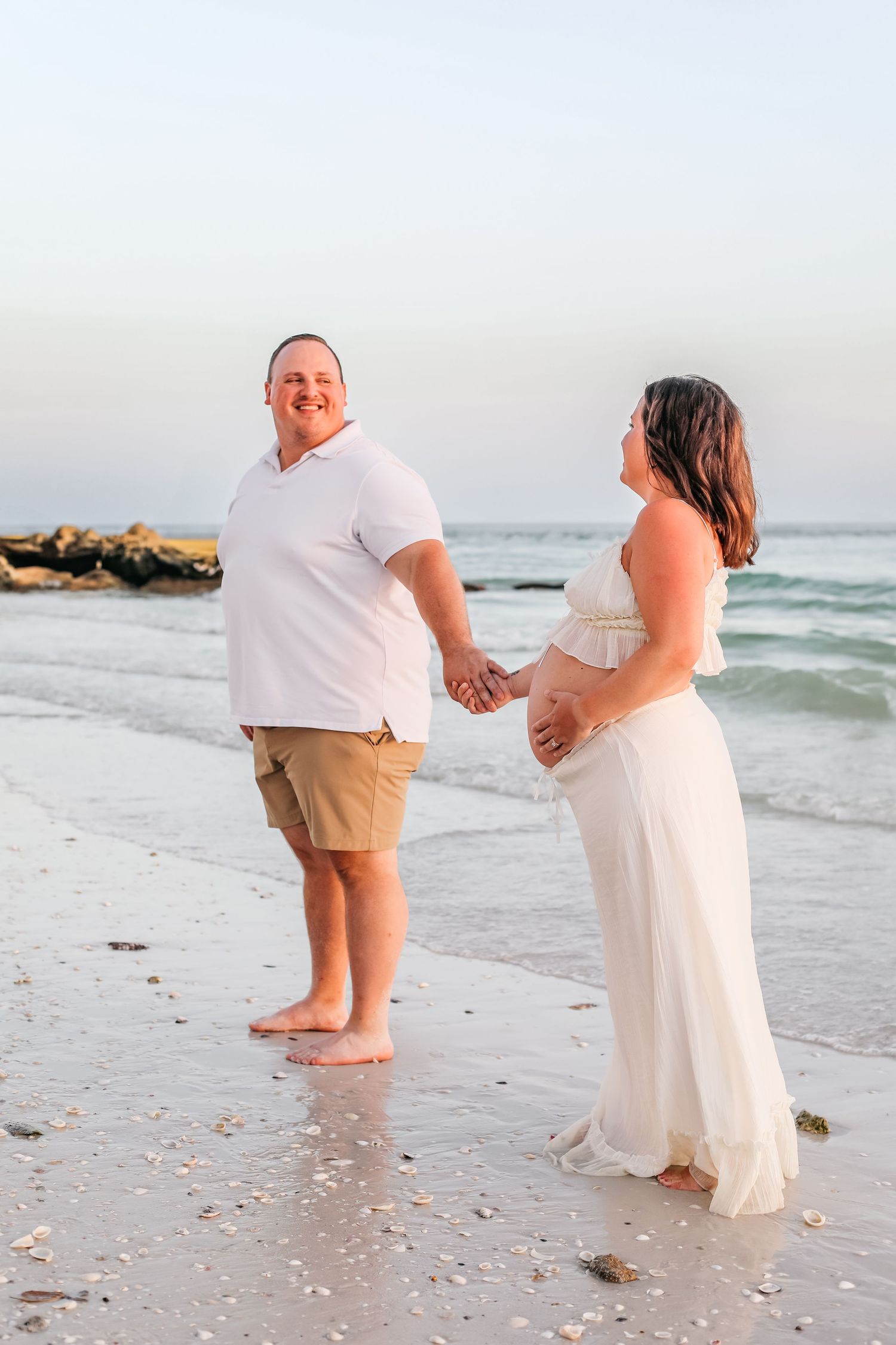 Couple holding hands while standing on beach during sunset maternity session.