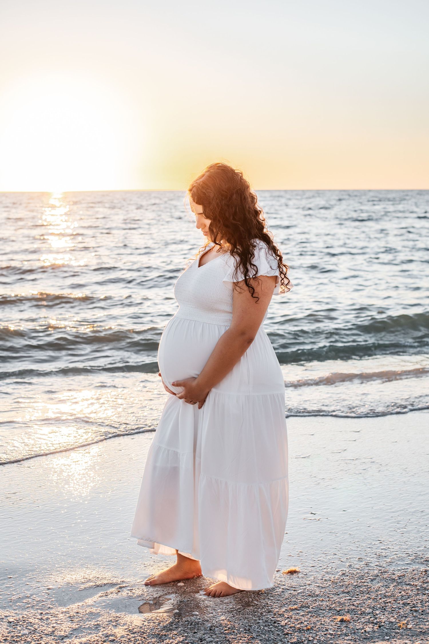 A figure in a flowing white dress walks along a sandy beach at sunset as waves roll onto the shore.