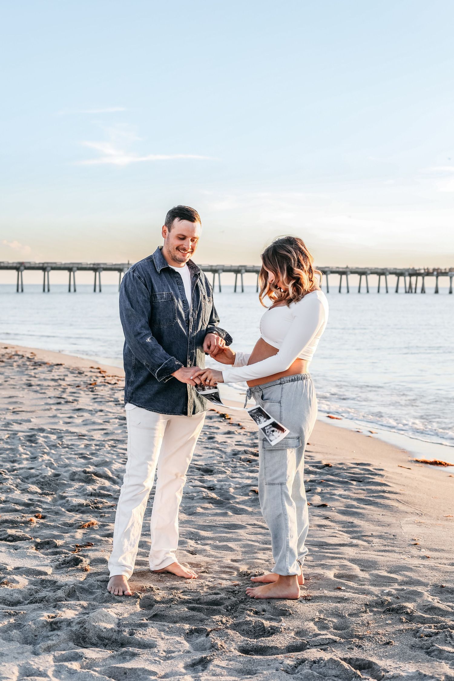 A couple shares a romantic moment on a sandy beach at sunset near a wooden pier.