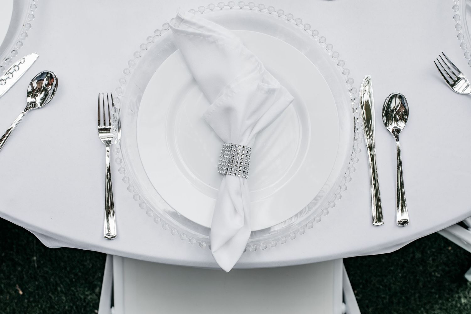 Elegant white place setting with folded napkin, rhinestone ring, and silver cutlery on a round table.