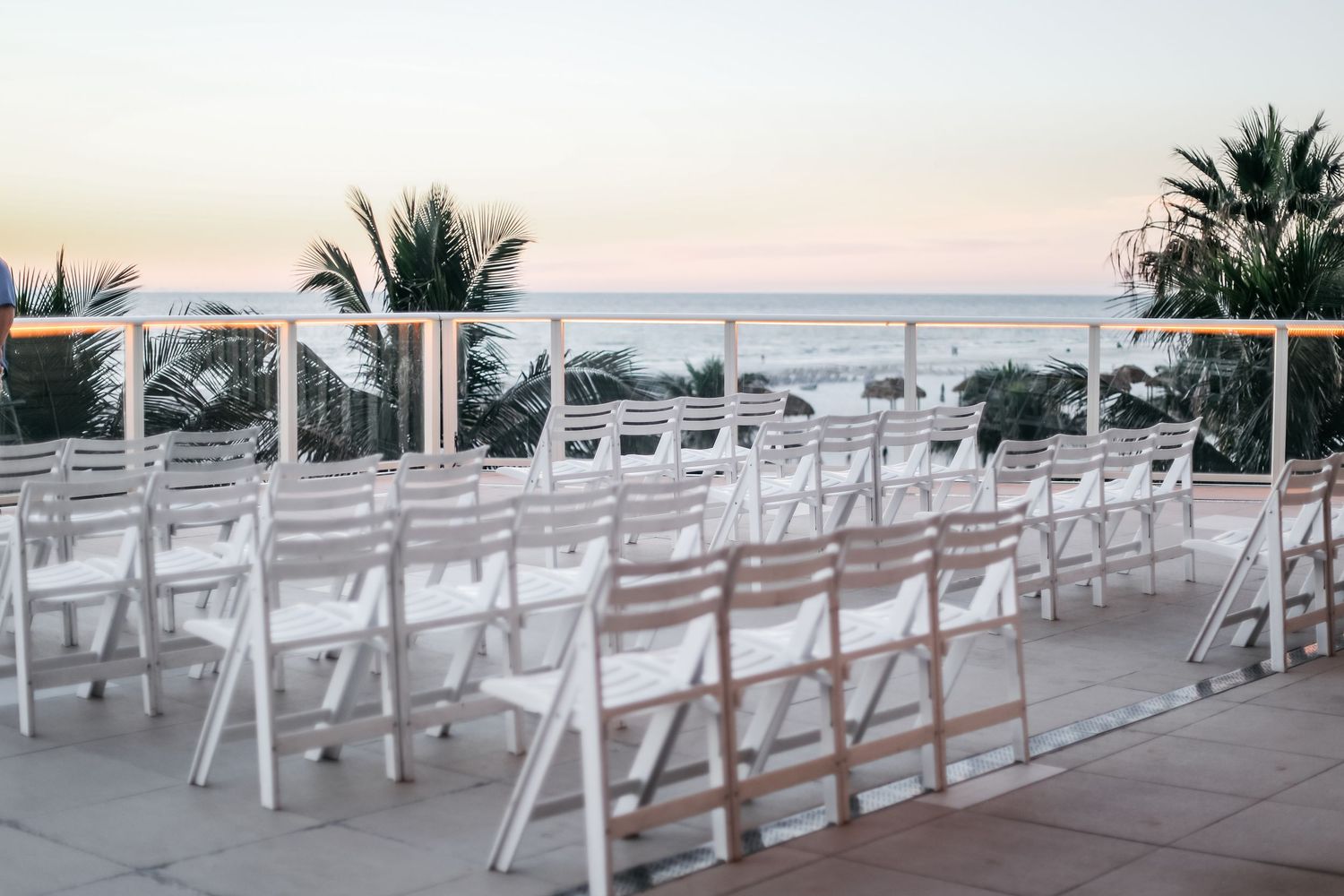 White folding chairs arranged for an outdoor beachside wedding ceremony at sunset with palm trees and ocean views.