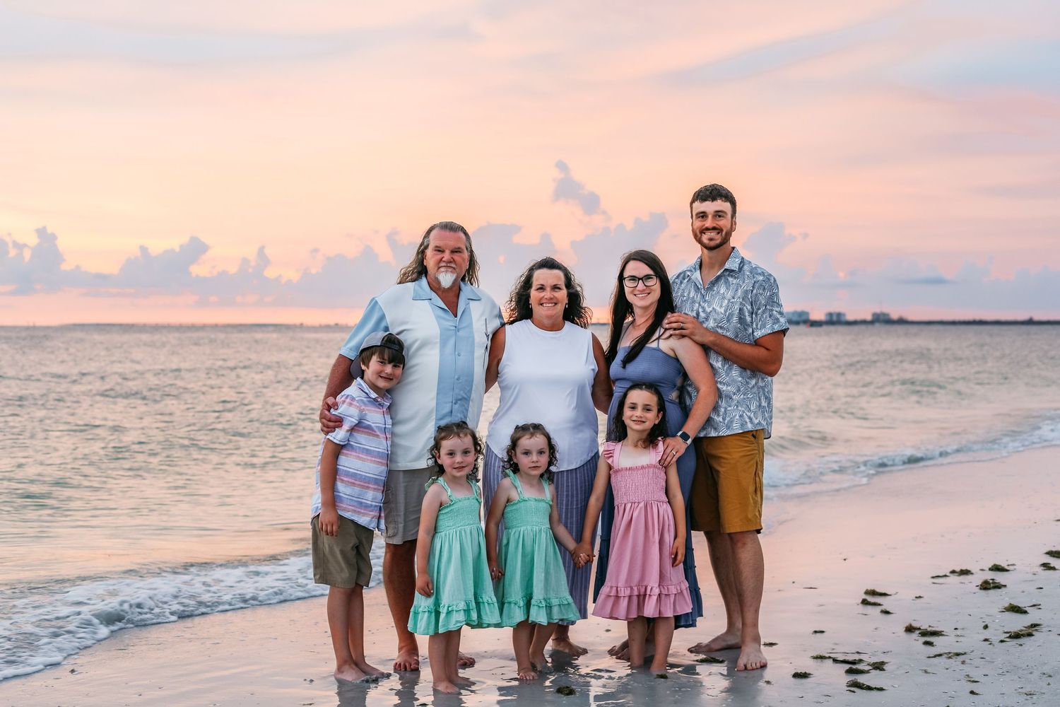 Family of seven poses on a beach at sunset with pink sky, waves, and children in colorful outfits.