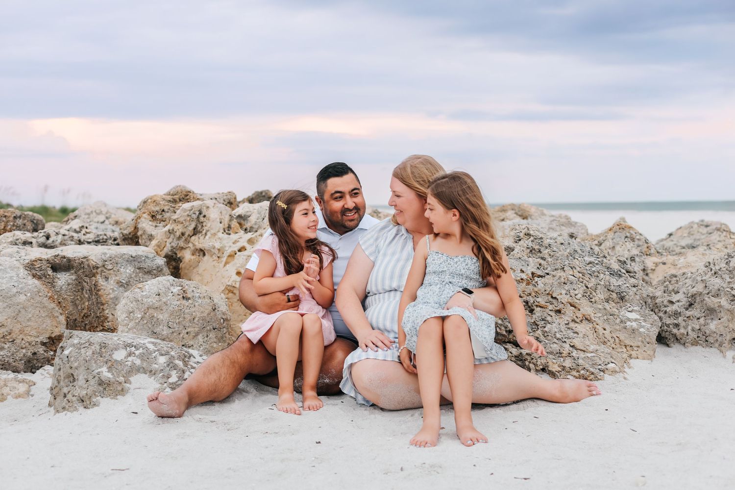 Family of four sitting together on sandy beach with rocky backdrop and pastel sunset sky.
