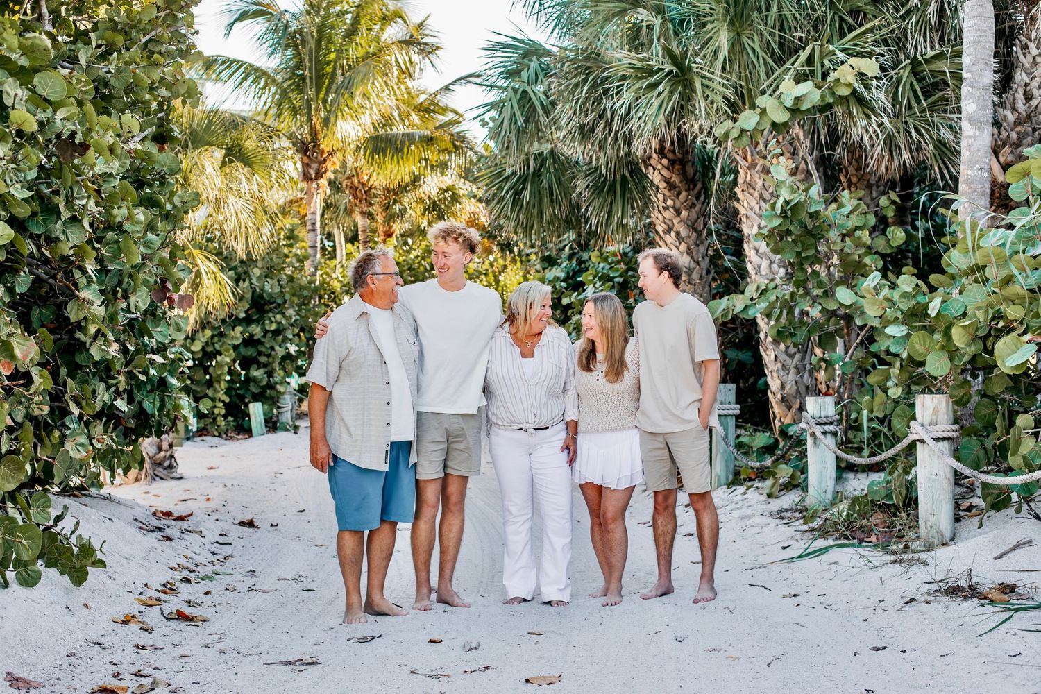 A group walking along a sandy beach path lined with tropical palm trees and lush vegetation in white and beige attire.