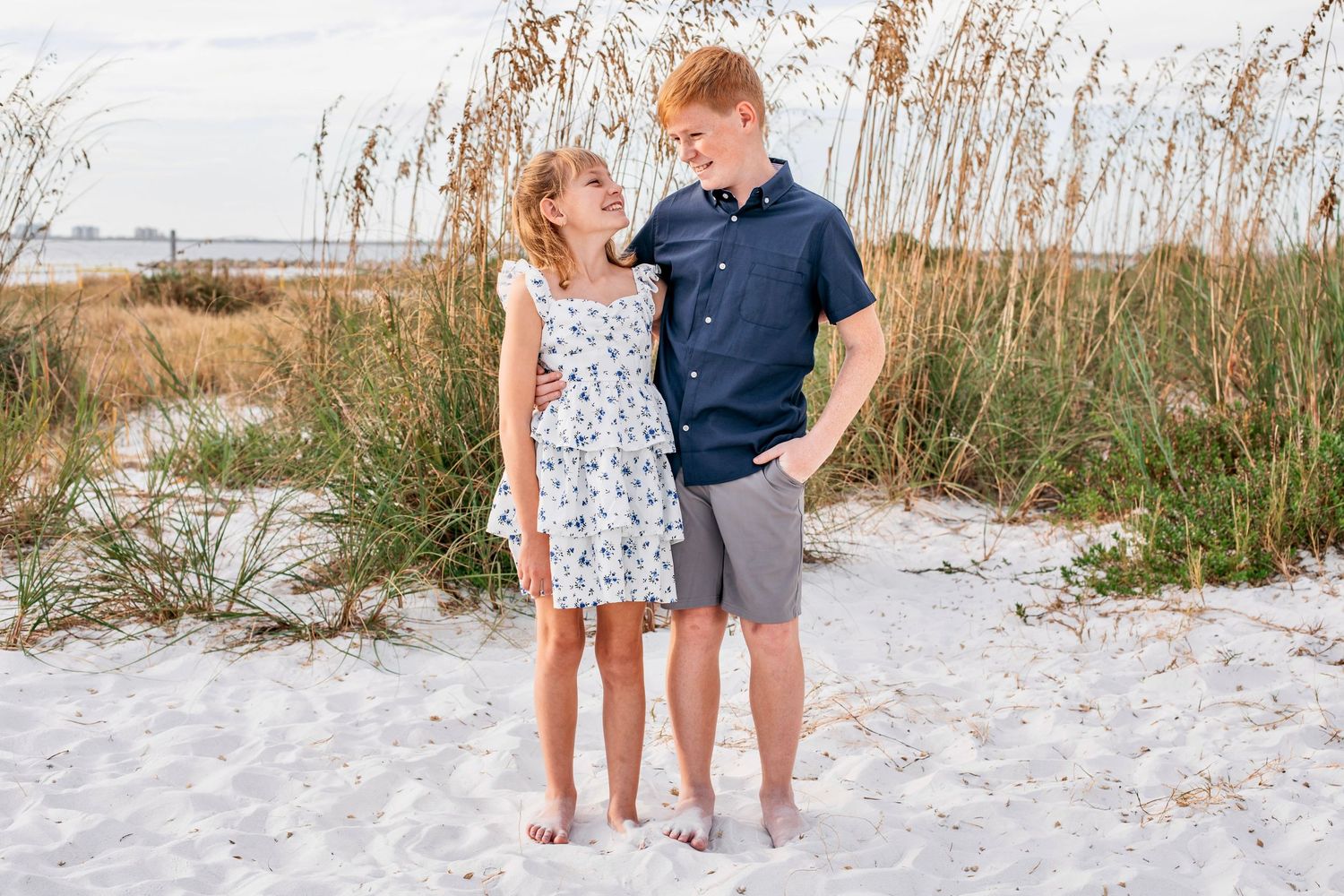 A couple stands together on a white sandy beach near tall sea oats during a beach photo session.
