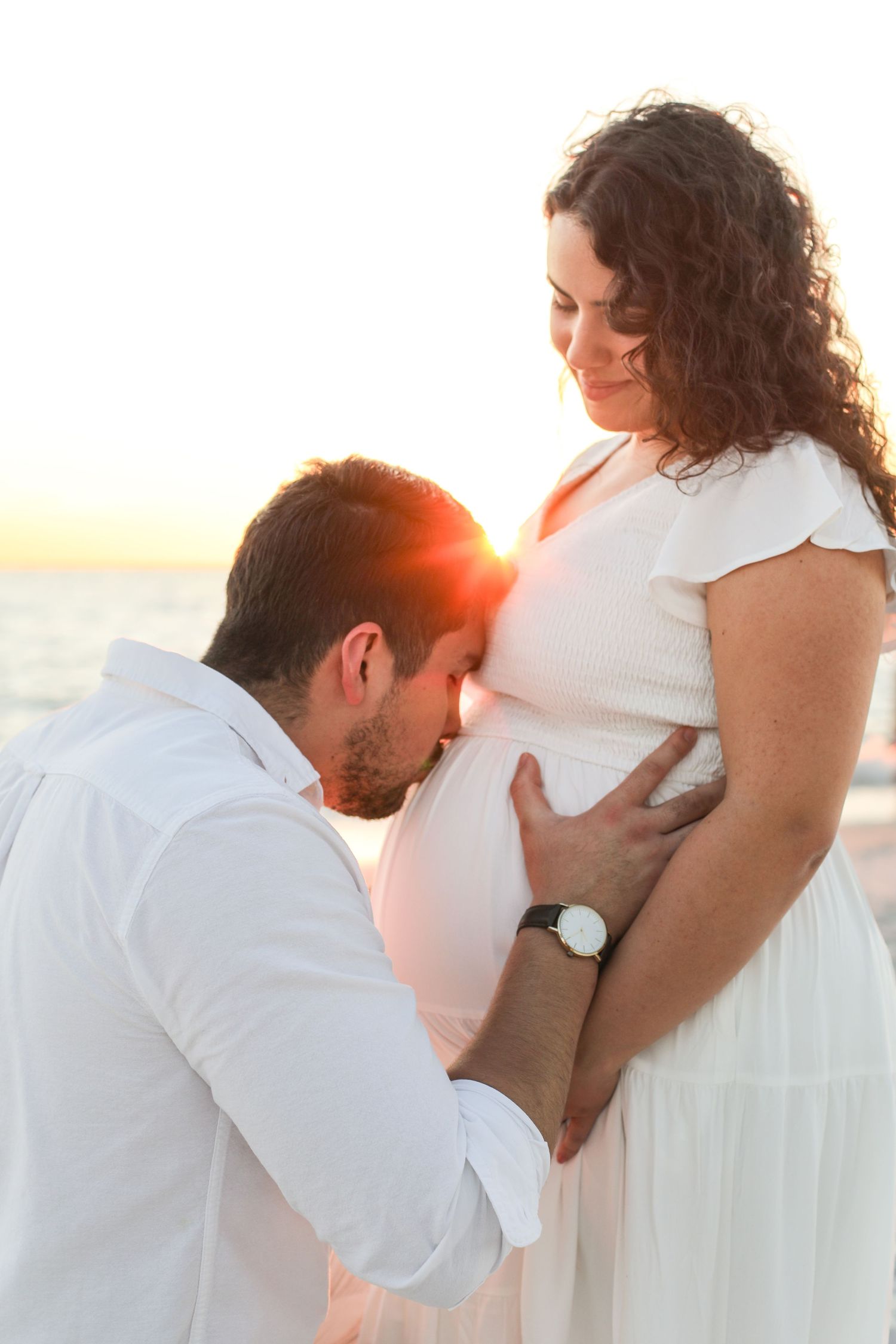 A couple in white clothing shares an intimate moment at sunset on the beach in a romantic photo series.