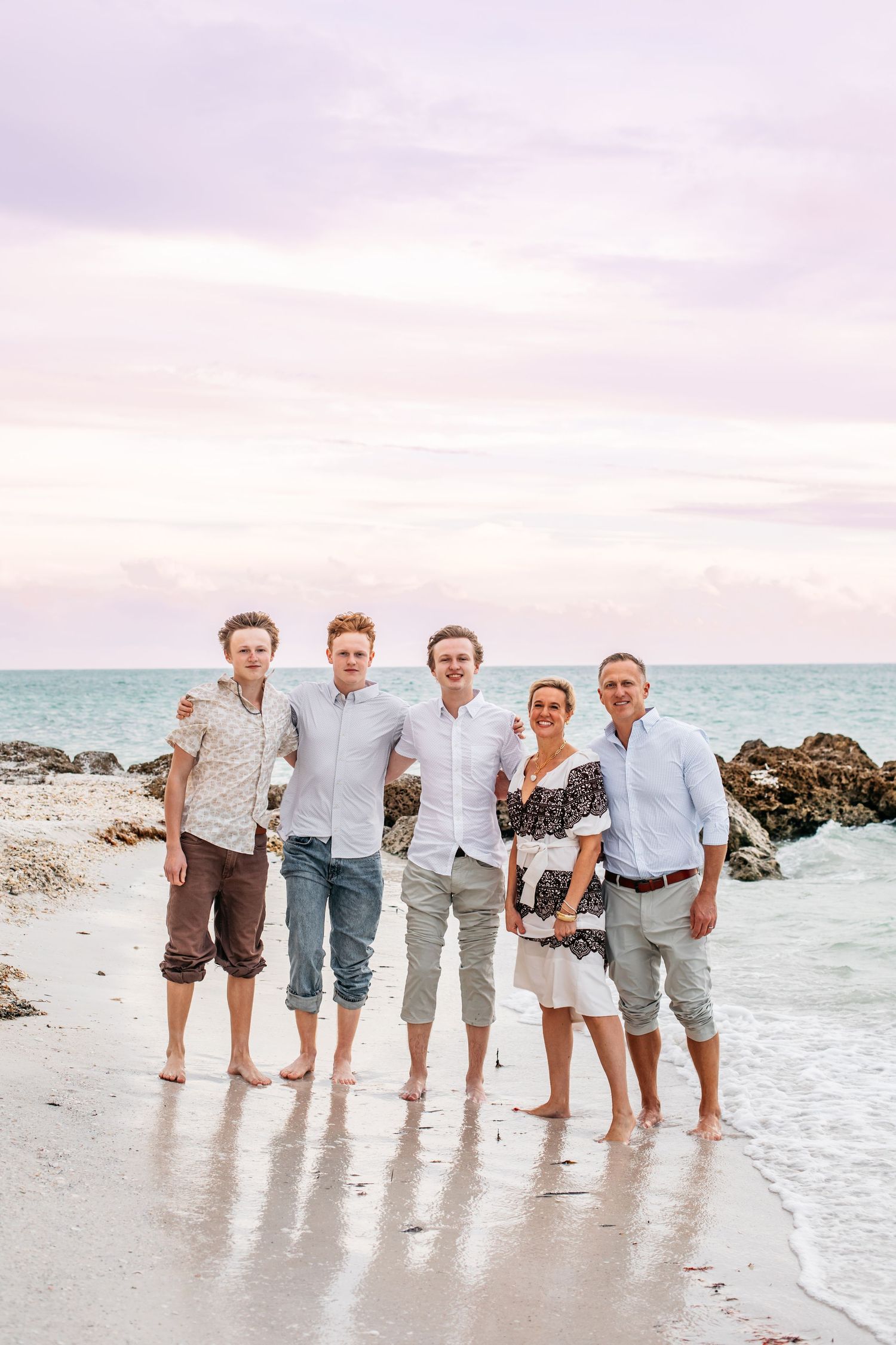 A group stands together at the water's edge on a beach during sunset, wearing casual summer clothing.