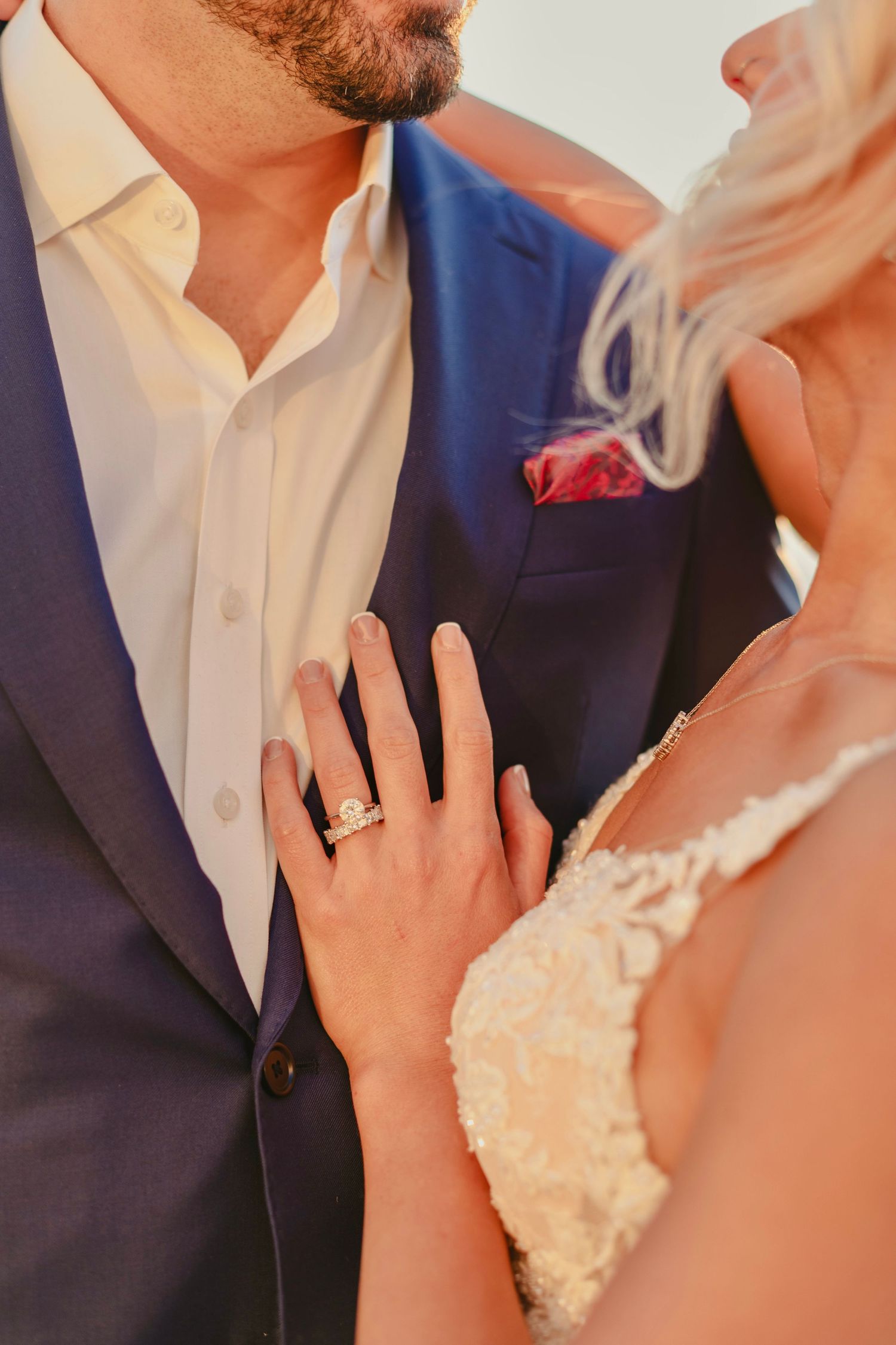 Bride in lace gown shows diamond engagement ring while holding groom in navy suit with red pocket square.