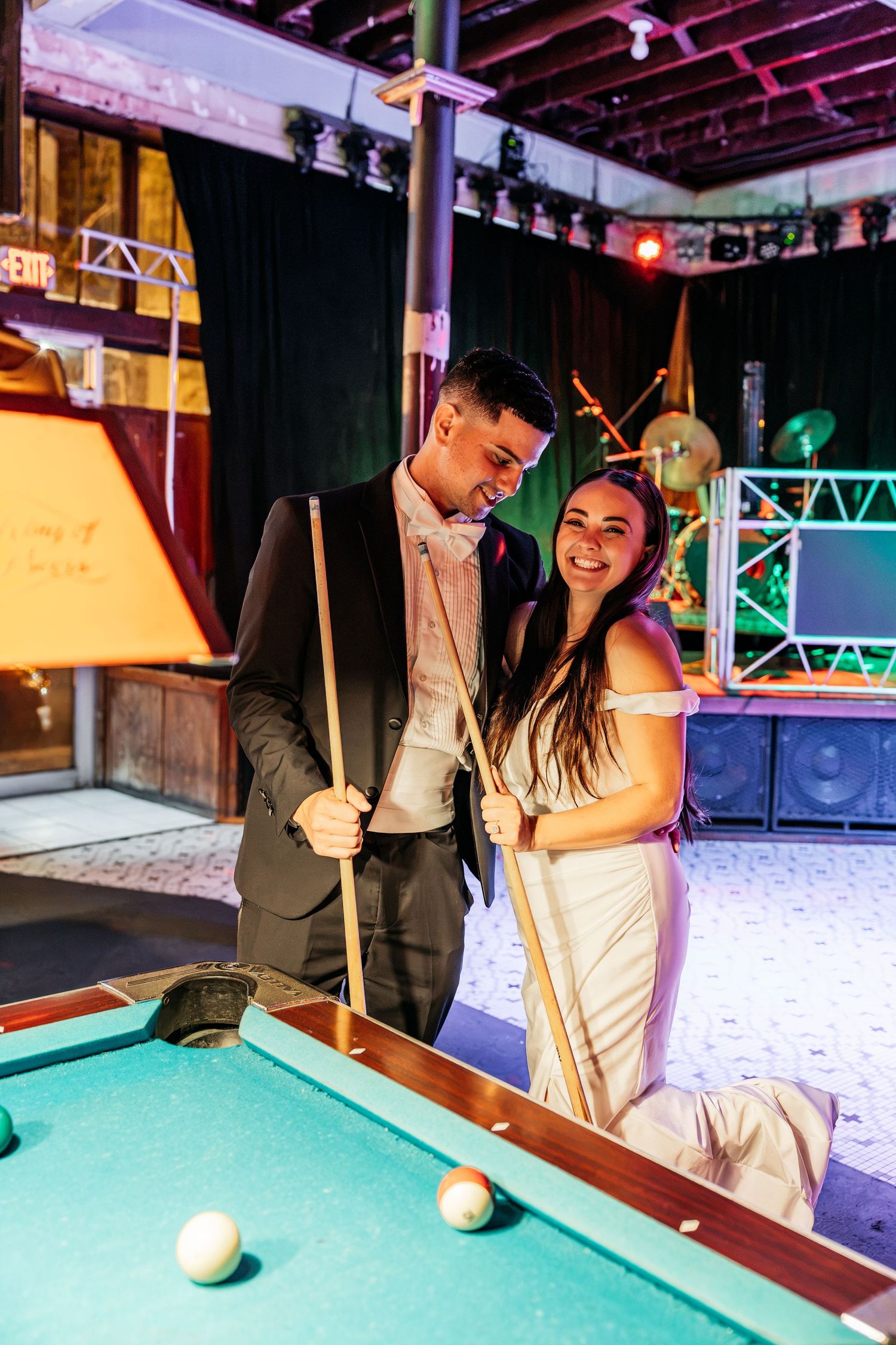 Bride and groom smiling together at a pool table, holding cue sticks at their wedding reception venue.