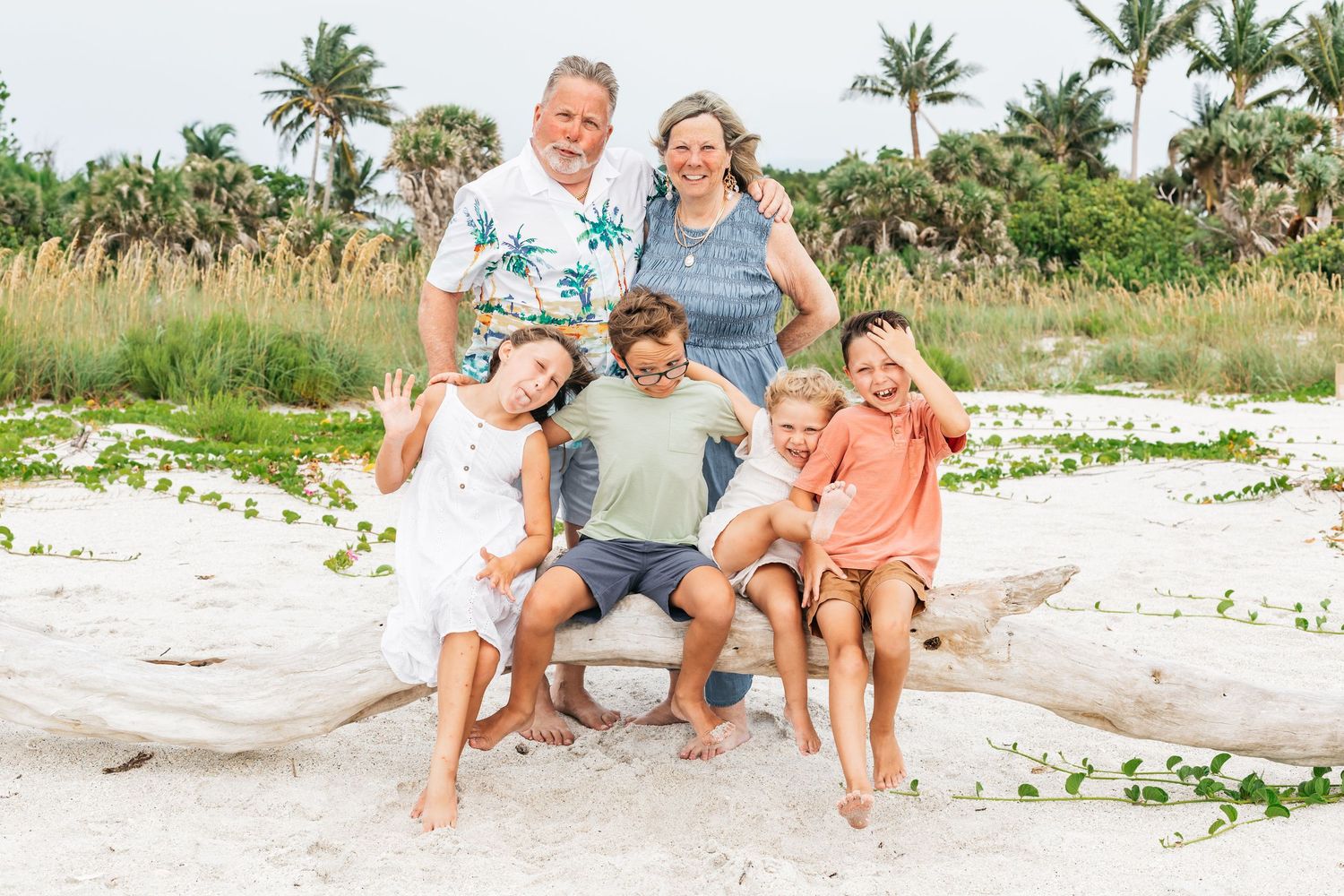 Family relaxing together on a white sandy beach with lush greenery and palm trees along the shoreline.