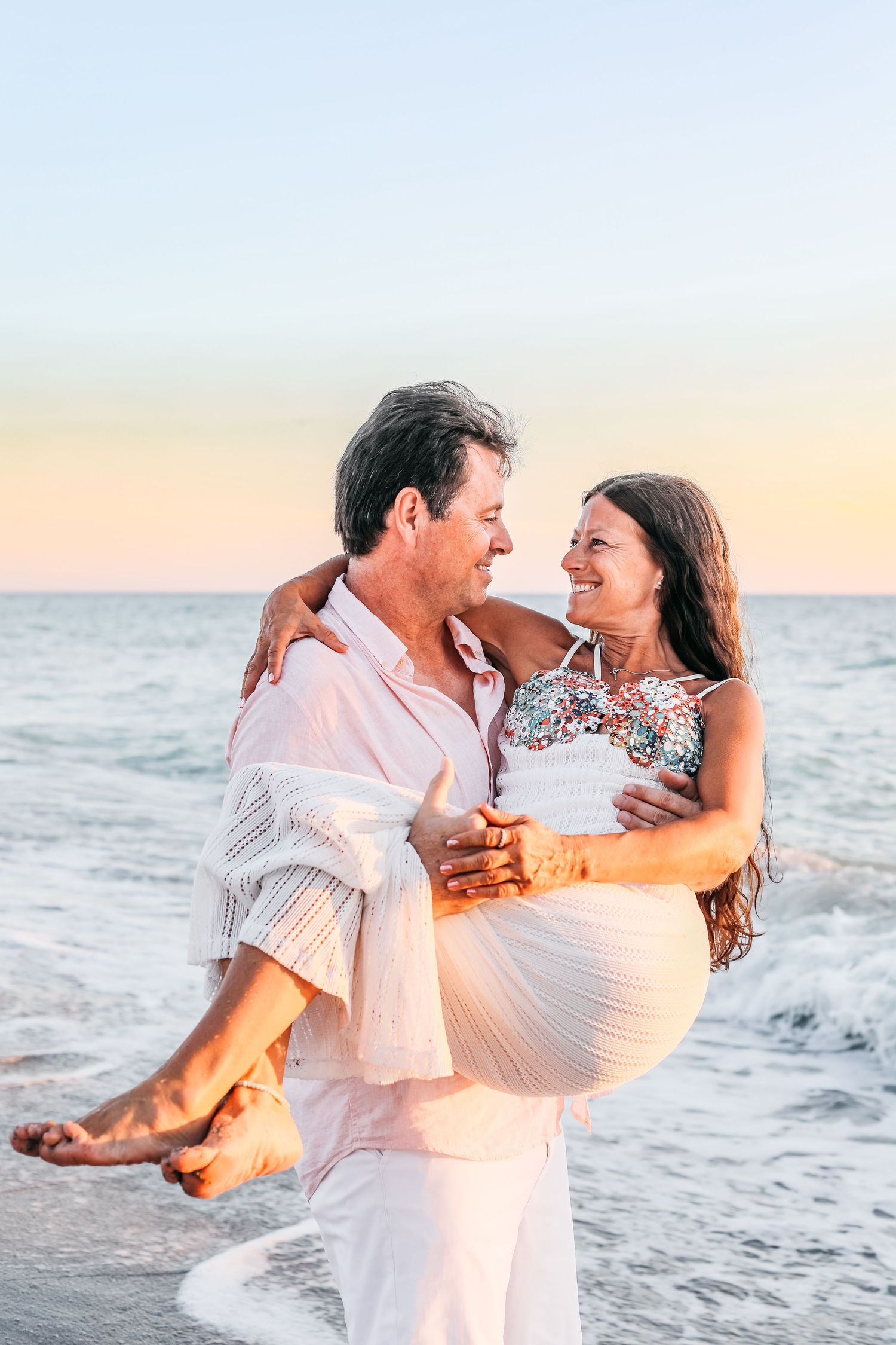 A romantic couple in white clothing dances and embraces on a beach at sunset with ocean waves in the background.