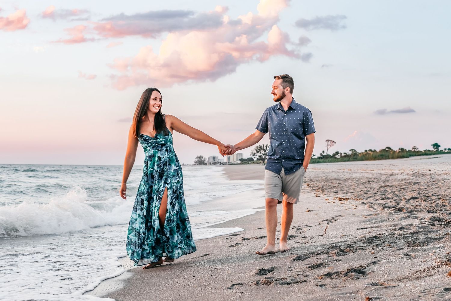 A couple walks hand in hand along a beach at sunset, with waves crashing and pink clouds in the sky.