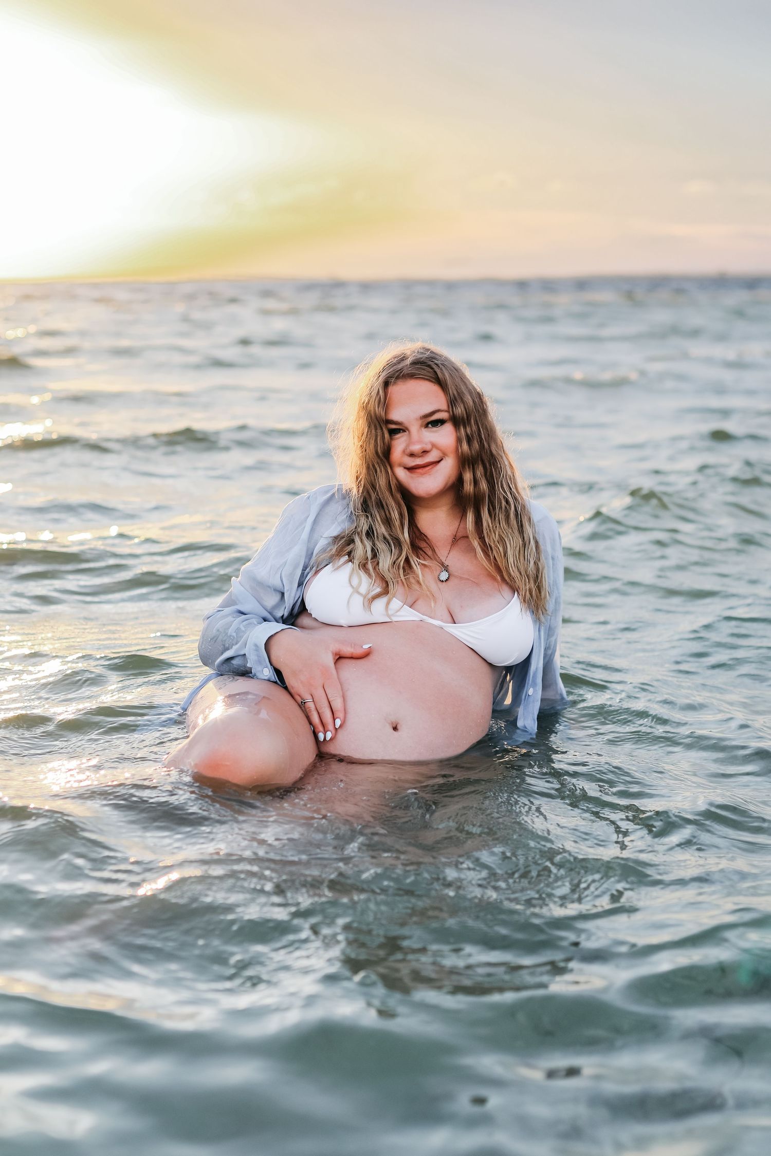 A person wearing a light colored top lies in shallow ocean water at sunset capturing beach maternity photos.
