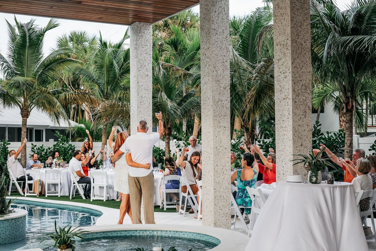 Outdoor tropical wedding reception by a pool with guests cheering as couple embraces under a pavilion surrounded by palm trees.